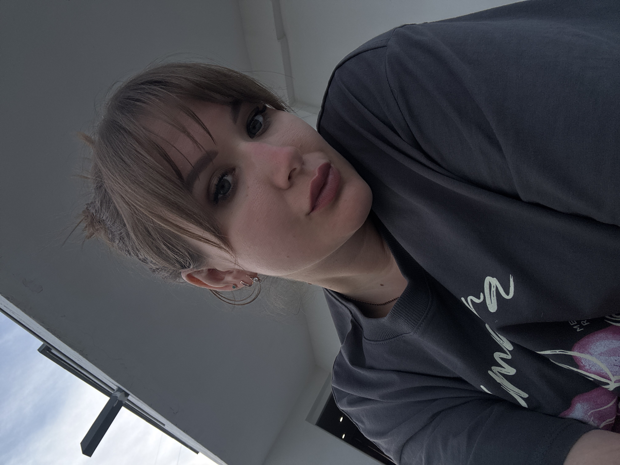 A young woman with light brown hair and bangs is pictured from a low angle. She has light skin and her lips are pursed slightly. She is wearing a gray t-shirt with white writing and a purple design. Her ear has multiple earrings.
The background consists of light gray surfaces, possibly the underside of a building or overhang. The sky with some clouds is visible in the lower left corner of the frame.
There is no information in the image to confirm it was taken in the city of Yevpatoriya, Ukraine.