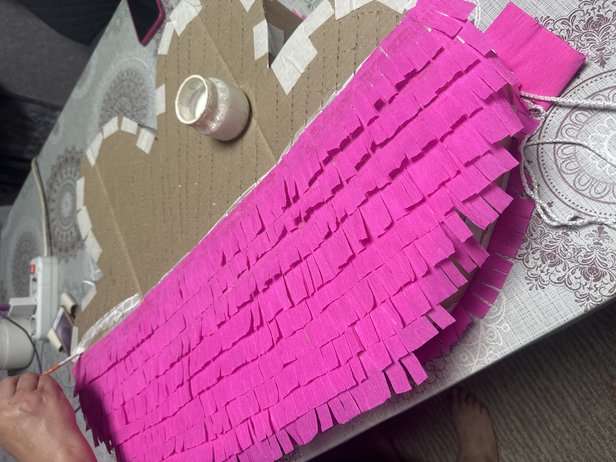 A partially completed piñata lies on a patterned table, presumably in a home in the city of Unknown, Unknown. The piñata is constructed from cardboard, reinforced with white tape and is partially covered in bright pink fringed crepe paper. A small jar of white glue sits nearby, along with a hot glue gun plugged into an outlet. A person’s hand holds a long, thin orange stick, possibly used for applying glue or manipulating the crepe paper. A white rope is attached to the piñata.