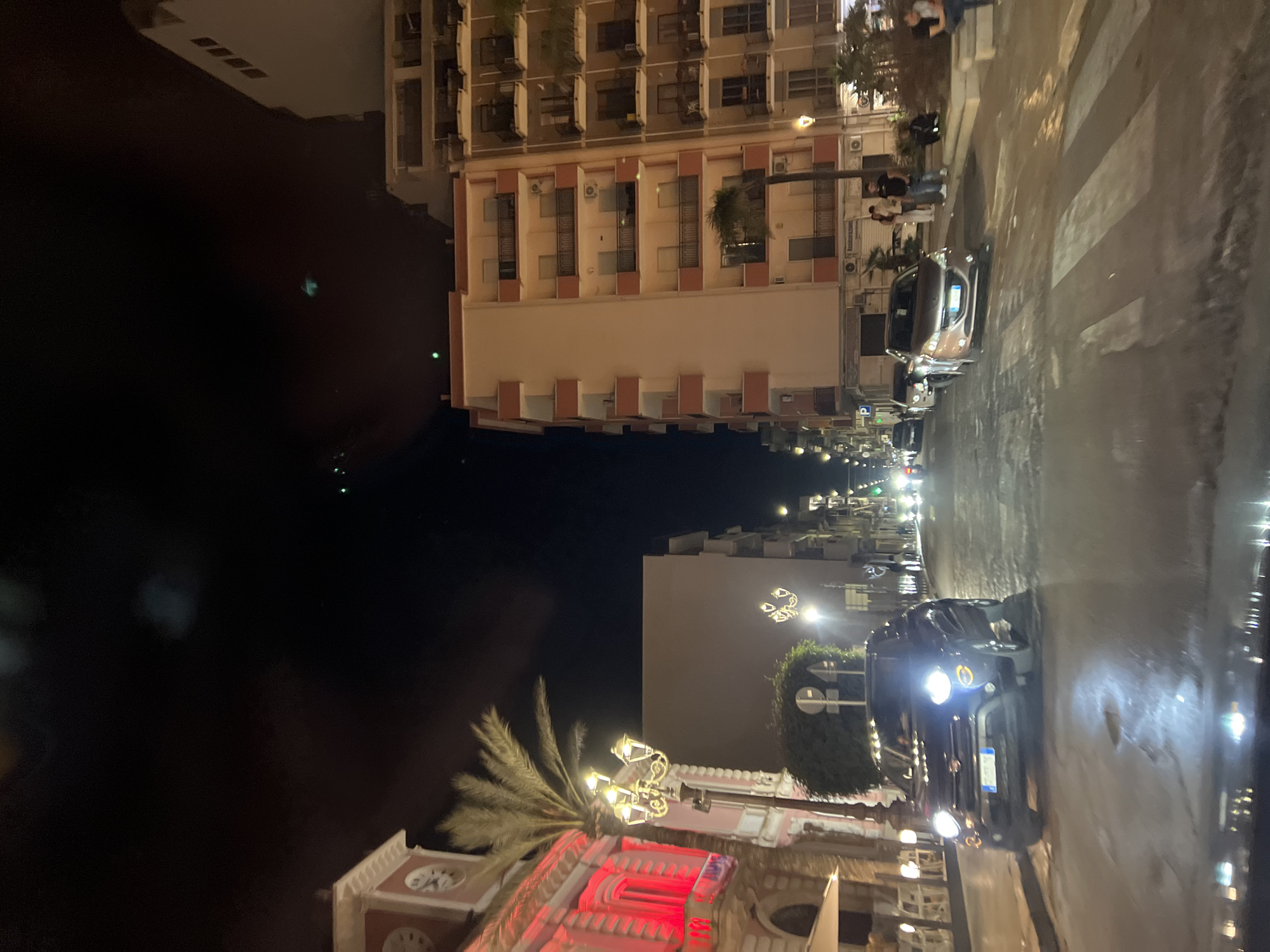 This is a night-time, eye-level, outdoor photograph taken from a vehicle looking down a street in Margherita di Savoia, Italy. The street is wet and reflects the lights of the buildings and vehicles. On the left side of the street is a building with a pink facade, illuminated with red lights, and a clock tower. A palm tree stands next to this building. Across the street, several multi-story apartment buildings are visible, some with balconies and air conditioning units. Streetlights with multiple lamps are present along the street. A dark-colored Fiat 500X is parked on the left side of the street, its headlights on. Several other cars are parked or driving on the street. A pedestrian crossing is visible in the foreground. Several people are standing on the sidewalk on the right side of the street. The sky is dark with some blurred lights.