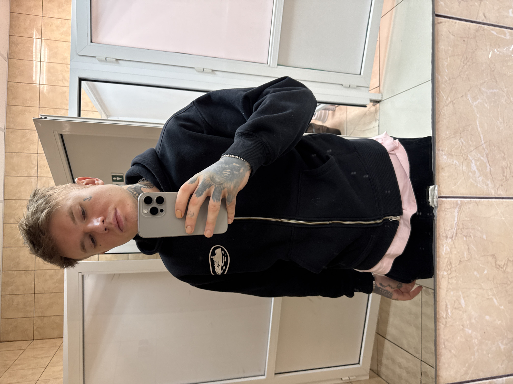 An indoor mirror selfie captures a young man, likely in his 20s or early 30s, in what appears to be a utilitarian indoor space in Moscow, Russia. He is looking directly into the camera lens of the silver/gray smartphone he holds with his right hand.
The man has short, light-colored hair and visible tattoos, including a small dark anchor or cross-like design below his right eye and extensive artwork covering his hands and forearms. He is wearing a black hooded zipper sweatshirt, under which a pink shirt is visible at the waist, and black trousers. A silver-colored chain bracelet is present on his left wrist. A small white circular logo with an indistinct stylized design is visible on the left side of his black sweatshirt.
The background, reflected in the mirror, consists of light beige tiled walls and plain white door frames and doors, characteristic of a restroom or changing room. The man maintains a neutral or slightly serious expression. The lighting suggests an indoor environment, likely illuminated by artificial light.