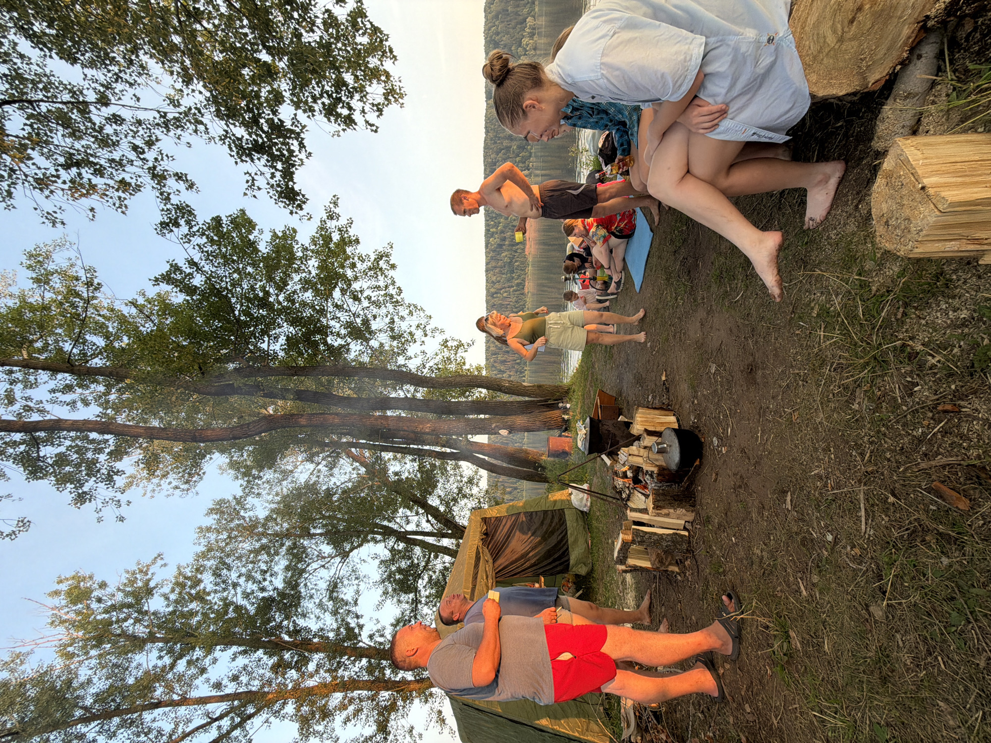 This image depicts an outdoor gathering of approximately ten individuals near a lake, likely in the city of Ol'khovskoye, Russia. The scene is set during the golden hour, with the setting sun casting a warm glow on the environment. In the foreground, a campfire is burning, surrounded by chopped wood. A large tent is pitched behind the campfire, suggesting a camping or outdoor event.
Several people are visible in the image. Two men are standing near the tent, one wearing a grey t-shirt and red shorts, the other in a blue t-shirt and light-colored shorts. A group of people is seated on the ground by the lake, some on mats. Among them, a woman in a green tank top and shorts stands, while a shirtless man in dark shorts stands nearby. Another woman in a light blue shirt is seated in the foreground, her legs crossed.
The environment includes tall trees with green foliage, a calm body of water, and a wooded hillside in the background. The ground is covered with dirt, grass, and scattered debris. The overall atmosphere suggests a relaxed social gathering and outdoor recreation.