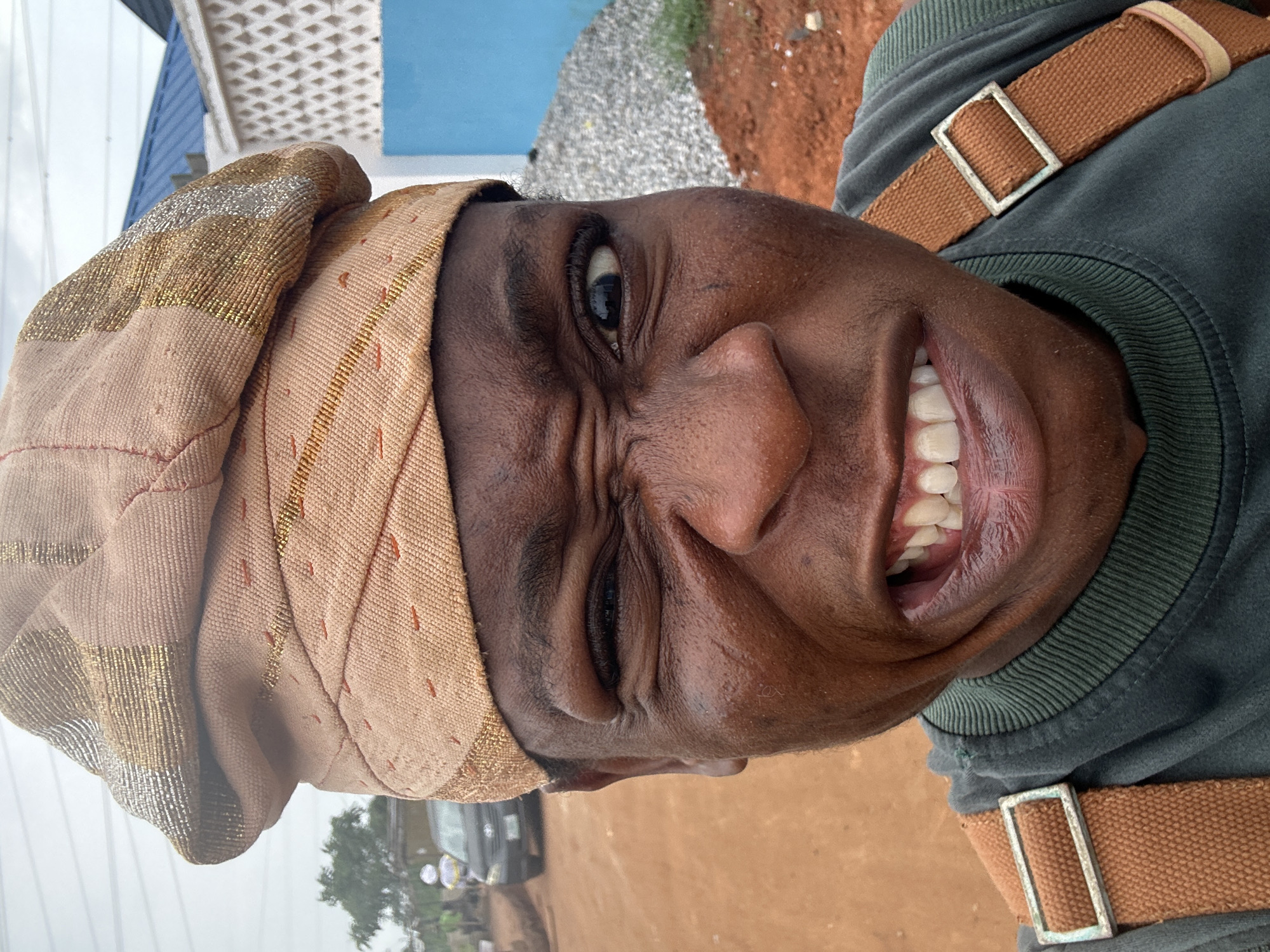 A person wearing a brown and gold head covering and a green shirt with brown straps is visible. The background includes a building with blue and white paneling, gravel, and red soil. In the distance, a car and trees are discernible. The person is making a facial expression. The image is rotated.