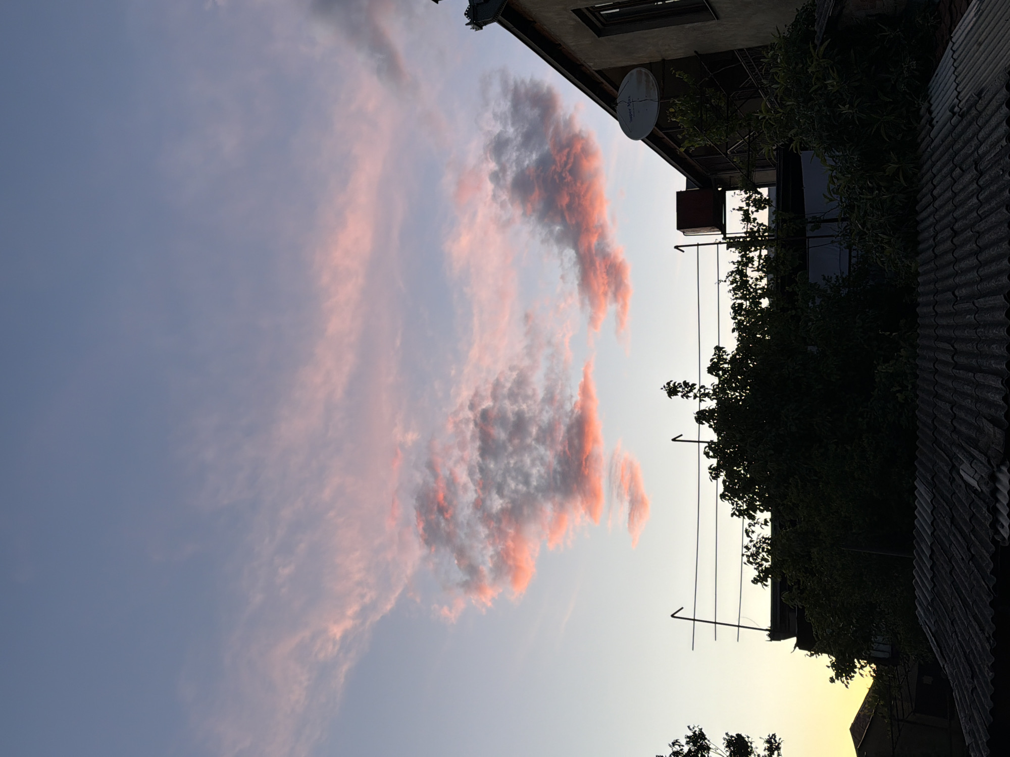 The image captures a scene at dusk or dawn in Tbilisi, Georgia. The sky is filled with light blue and pink-tinged clouds. A building is visible on the right side of the frame, featuring a satellite dish, foliage, and a corrugated roof. An antenna is also visible on the building. The angle suggests the photograph was taken looking upwards.