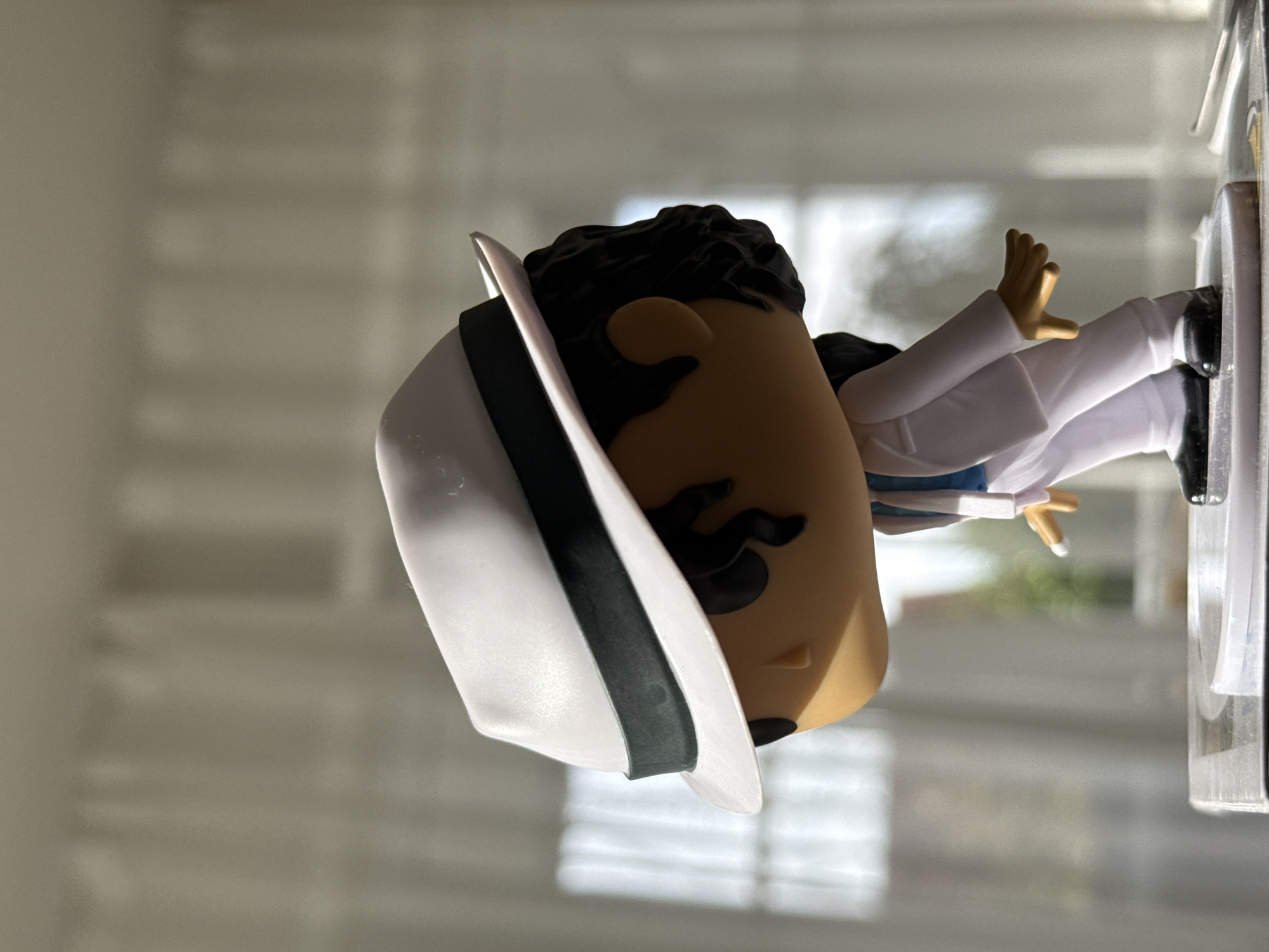 A close-up, profile view shows a Michael Jackson Funko Pop figure. The figure is wearing a white fedora with a black band, a white suit jacket with blue trim, white pants, and black shoes. The figure is positioned on a clear plastic stand, which is placed on a surface that appears to be a book or display box. The background consists of softly lit sheer curtains, with indistinct shapes of windows and possibly outdoor greenery visible through them, suggesting an indoor setting with natural light. There is no indication of the location being in Bad Sachsa, Germany.