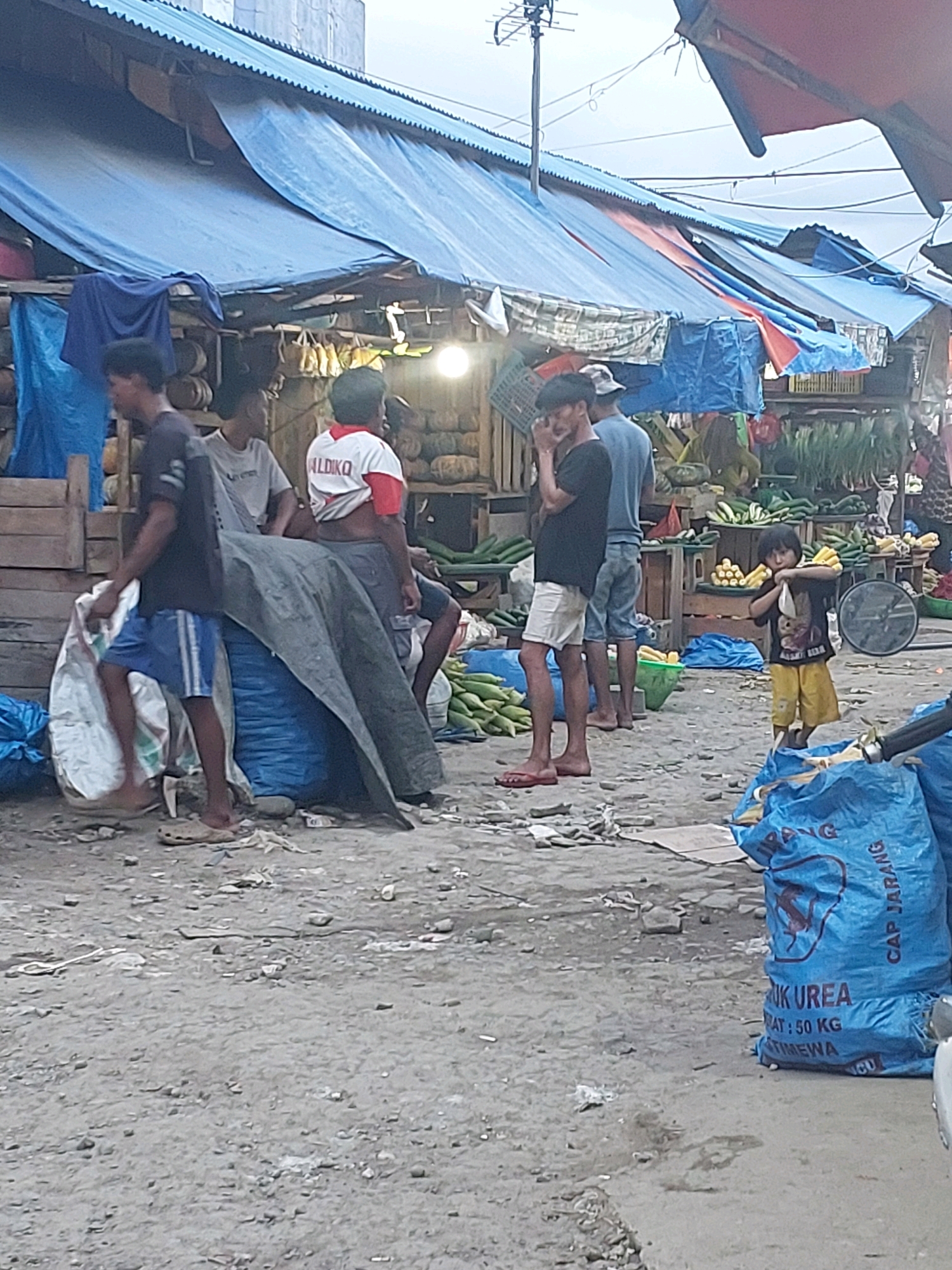 The image depicts an outdoor market scene in Ambon, Indonesia. In the foreground, a person in a black t-shirt and white shorts is walking to the left, wearing red flip-flops. To their right, a large blue sack is visible, partially obscuring other elements. Behind the person, a group of individuals are gathered near market stalls. One person is wearing a white t-shirt with red trim and the word "ALDIKO" across the chest, grey shorts, and is looking to the left. Another person, in a grey t-shirt, is seated and looking to the right. A third person, in a blue t-shirt and shorts, is standing and facing away from the camera.
The market stalls are covered with blue tarps, some of which are partially pulled back, revealing goods such as pumpkins and other produce. The ground is uneven and appears to be made of dirt and gravel. In the mid-ground, several stacks of corn are visible, and a young child in a black t-shirt and yellow shorts is holding a bundle of corn. Further back, more produce, including cucumbers, is displayed on wooden stands. The overall impression is of a bustling, informal market environment. The sky above is overcast, suggesting a cloudy day.