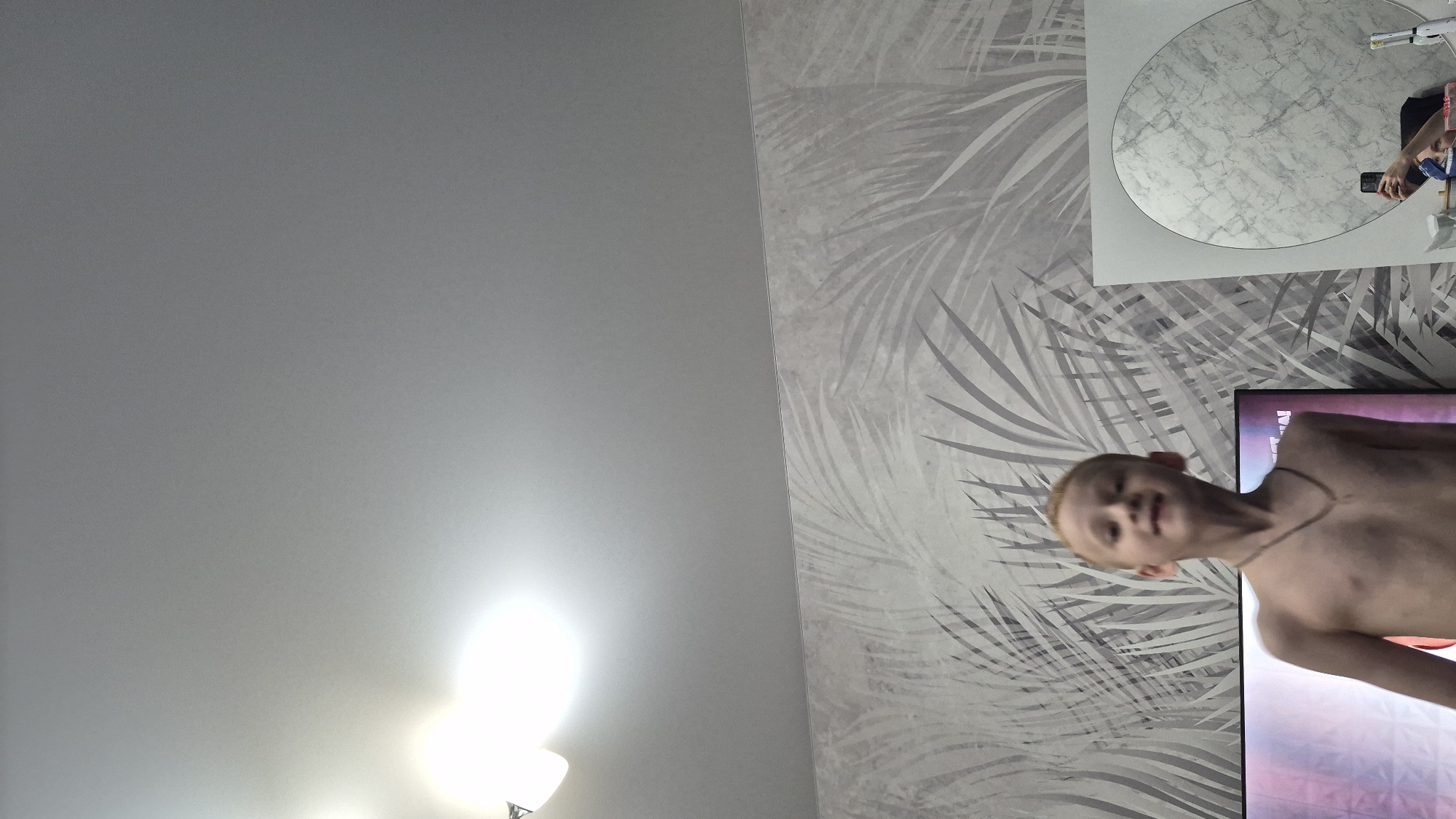 The image is a photograph taken indoors, rotated 90 degrees clockwise. It captures a domestic scene featuring a young boy, a mirror, and a television, set against a backdrop of a patterned wall and a plain ceiling.
The left half of the image (which would be the ceiling in an upright view) is a large, plain light-grey surface. In the bottom-left corner of this section, there is a very bright, diffused light source, possibly a lamp or window, casting a strong glare.
The right half of the image (which would be a wall in an upright view) is covered in wallpaper featuring a repeating pattern of stylized palm leaves in shades of light grey and white.
Mounted on this patterned wall, in the upper-right quadrant of the image, is a circular mirror with a thin white frame. The mirror's reflective surface has a light grey, mottled, marble-like appearance. Reflected in the mirror is a person holding a black smartphone, seemingly taking the photograph. A hand, part of an arm, and a blue and white patterned garment are visible in the reflection.
In the lower-right quadrant of the image, a young boy is visible from the chest up. He has light-colored hair and is wearing a thin, dark necklace. He is shirtless and appears to be looking towards the camera with a neutral to slightly smiling expression. His head is slightly tilted.
Behind and partially below the boy, a large, rectangular screen, likely a television, is visible. The screen displays colorful content with hues of purple, red, and possibly other colors, suggesting a video or program is playing.
The overall scene depicts a casual moment in a room, possibly a living area or bedroom, with bright indoor lighting. No specific text is visible in the image.