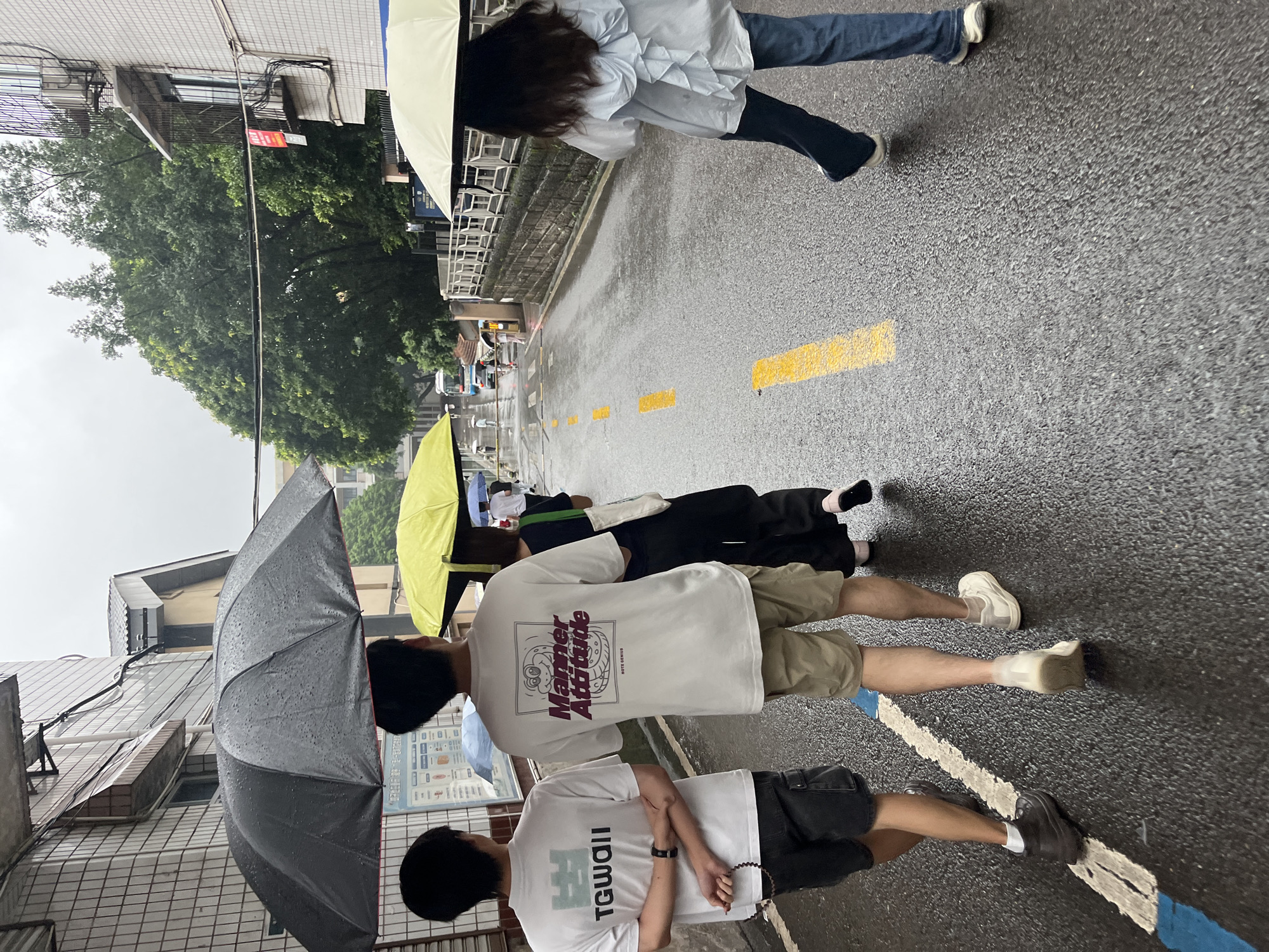 The image captures a rainy day scene on a wet asphalt street in an urban area, identified as Yuzhong District, China. The road is dark and reflective, indicating recent or ongoing precipitation, with visible water droplets on the surface and umbrellas.
In the foreground, a group of three individuals walks away from the camera. The leftmost person, a male, wears a white short-sleeved T-shirt with a green graphic reading "TGWAI!" on the back, black cargo shorts, dark shoes, and white socks. He has a dark beaded bracelet on his left wrist and his arms are crossed at the back. To his right, a second male wears a white short-sleeved T-shirt featuring a purple "Manner Attitude" graphic with a cartoon worm-like character and "NOTE GENIUS" text, khaki shorts, and white sneakers. Both males are partially covered by a shared large black umbrella, visibly wet with raindrops. To the right of the second male, a third person, likely female, is mostly obscured by a bright yellow umbrella; her lower body shows dark trousers and pink flat shoes.
Further ahead on the right side of the street, a fourth person, a female, walks independently. She wears a light blue long-sleeved shirt with a prominent ruffled bow detail on the back, blue flared jeans, and white sneakers. She holds a light cream-colored umbrella over herself.
The street itself is marked with a white solid line on the left, a short blue line alongside it in the foreground, and yellow dashed lines running down the center. Buildings flank both sides of the street. On the left, a multi-story building with a light-tiled exterior features exposed electrical wiring and a partially visible informational board. On the right, another multi-story building with a similar facade includes a balcony with a metal railing, an air conditioning unit, and a red sign with Chinese characters. A stone retaining wall with a metal railing runs along the right side of the street further into the background. Dense green trees are visible above and between the buildings. In the distance, several other pedestrians are visible, most carrying umbrellas, and a blue and white bus is parked or moving on the left side of the road. The sky is overcast, consistent with rainy weather.