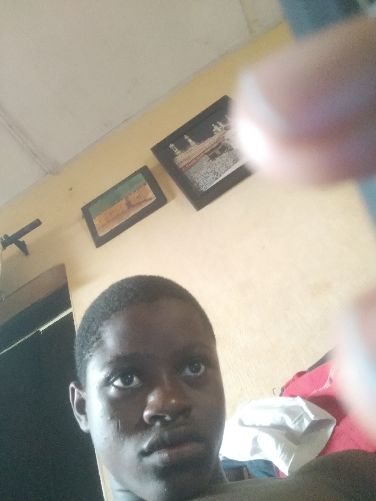 A close-up, slightly upward-angled image captures a young, dark-skinned person, likely male, with short dark hair, looking towards the camera. Their expression is neutral and calm. The upper right portion of the photograph is blurred by what appears to be fingers or a thumb, indicating the image is a selfie.
The setting is an indoor room with light yellow or beige walls and a white ceiling. On the left side of the frame, a dark structure, possibly a door or cabinet, is visible. A dark-colored ceiling fan is mounted on the wall to the left. Two framed pictures hang on the wall. The smaller frame on the left displays an image of light-colored buildings in what appears to be a desert or arid landscape. The larger frame on the right features a detailed architectural scene, potentially a cityscape or a religious site like the Kaaba, often seen in Islamic homes. In the lower right background, a pile of folded clothes or fabric, including red and white items, can be seen. The lighting is diffused, consistent with an indoor setting during the day or under artificial light. No discernible text is visible. The scene suggests a personal, everyday environment, typical of a home in Shangisha, Nigeria.