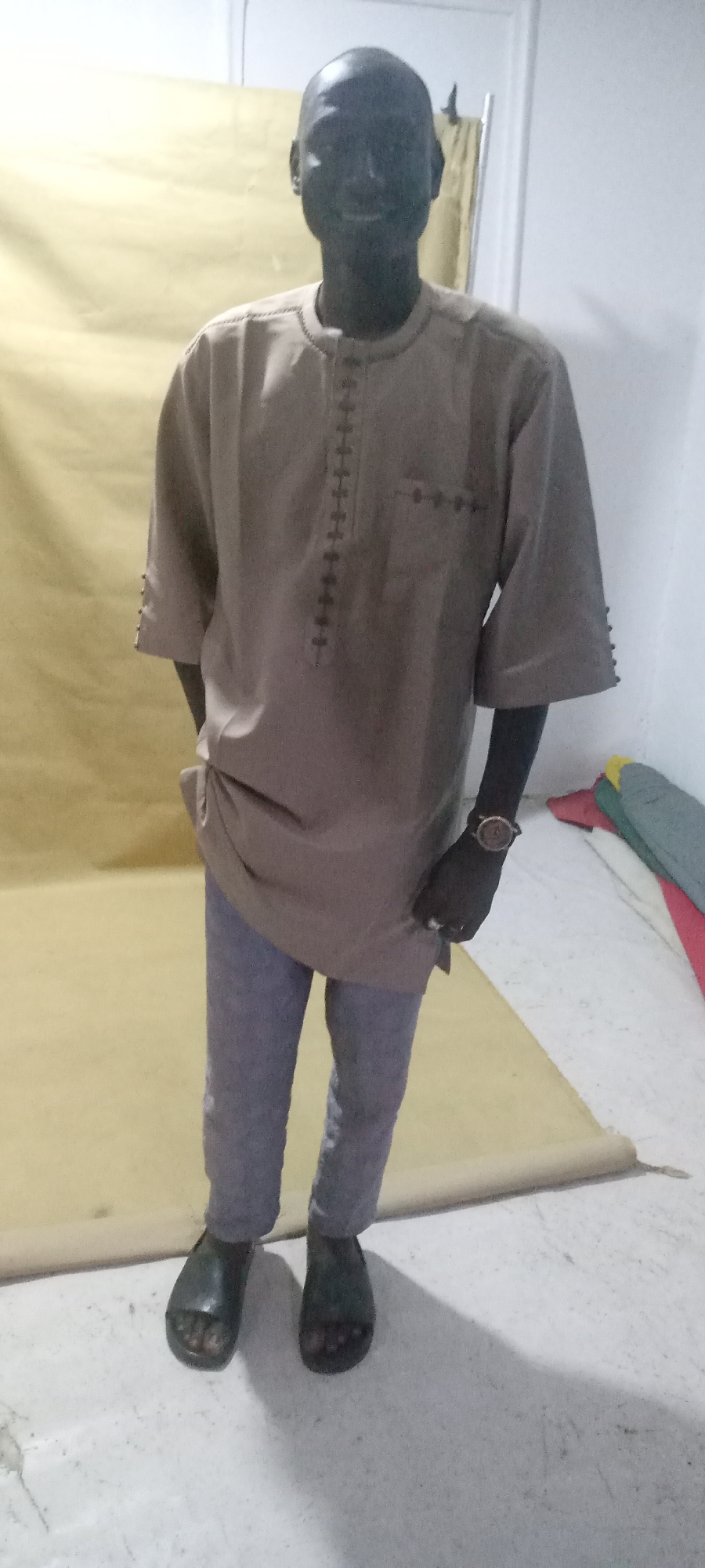 A man stands indoors against a yellow backdrop. He is wearing a beige shirt with embroidered detailing and grey pants. He is also wearing black sandals. The lighting is somewhat dim. The location of the image is Jos, Nigeria.