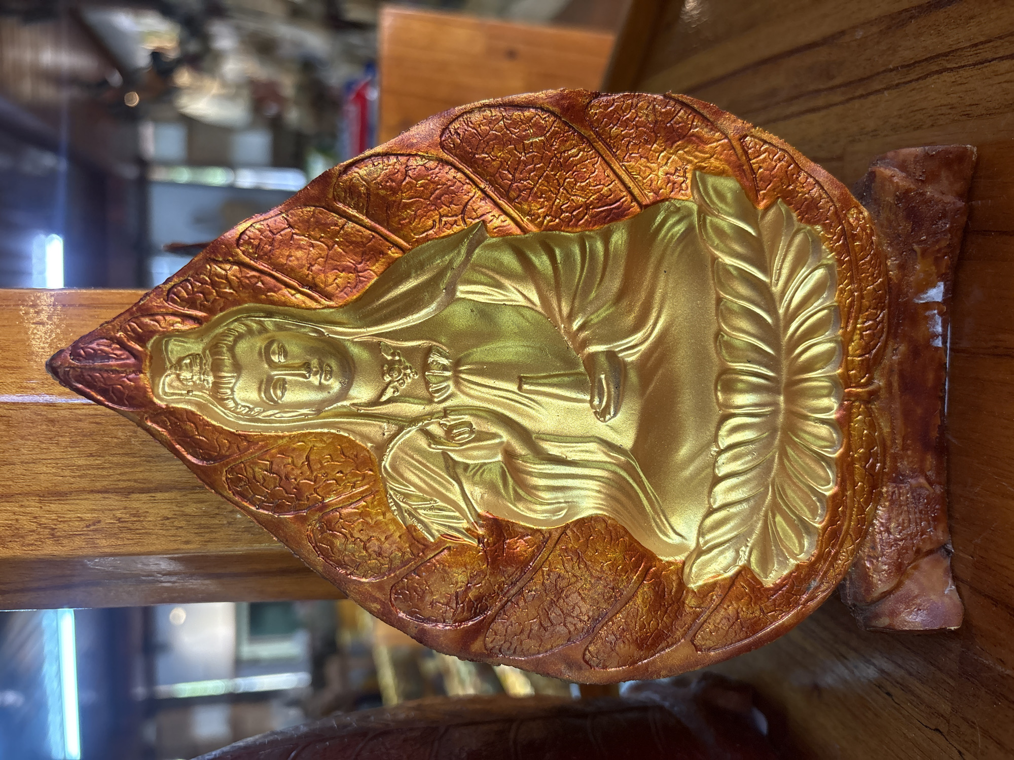 The image shows a golden statue of a deity, likely Guan Yin, in a leaf-shaped frame with a textured orange and gold finish. The statue is seated on a lotus pedestal. The background is out of focus and appears to be an indoor setting with wooden elements and fluorescent lighting. The statue is positioned on a wooden surface, possibly a table or shelf. There are no people or visible actions in the image. The overall impression is of a devotional or decorative item. The image does not contain any information that would suggest a specific location in Đà Nẵng, Vietnam.