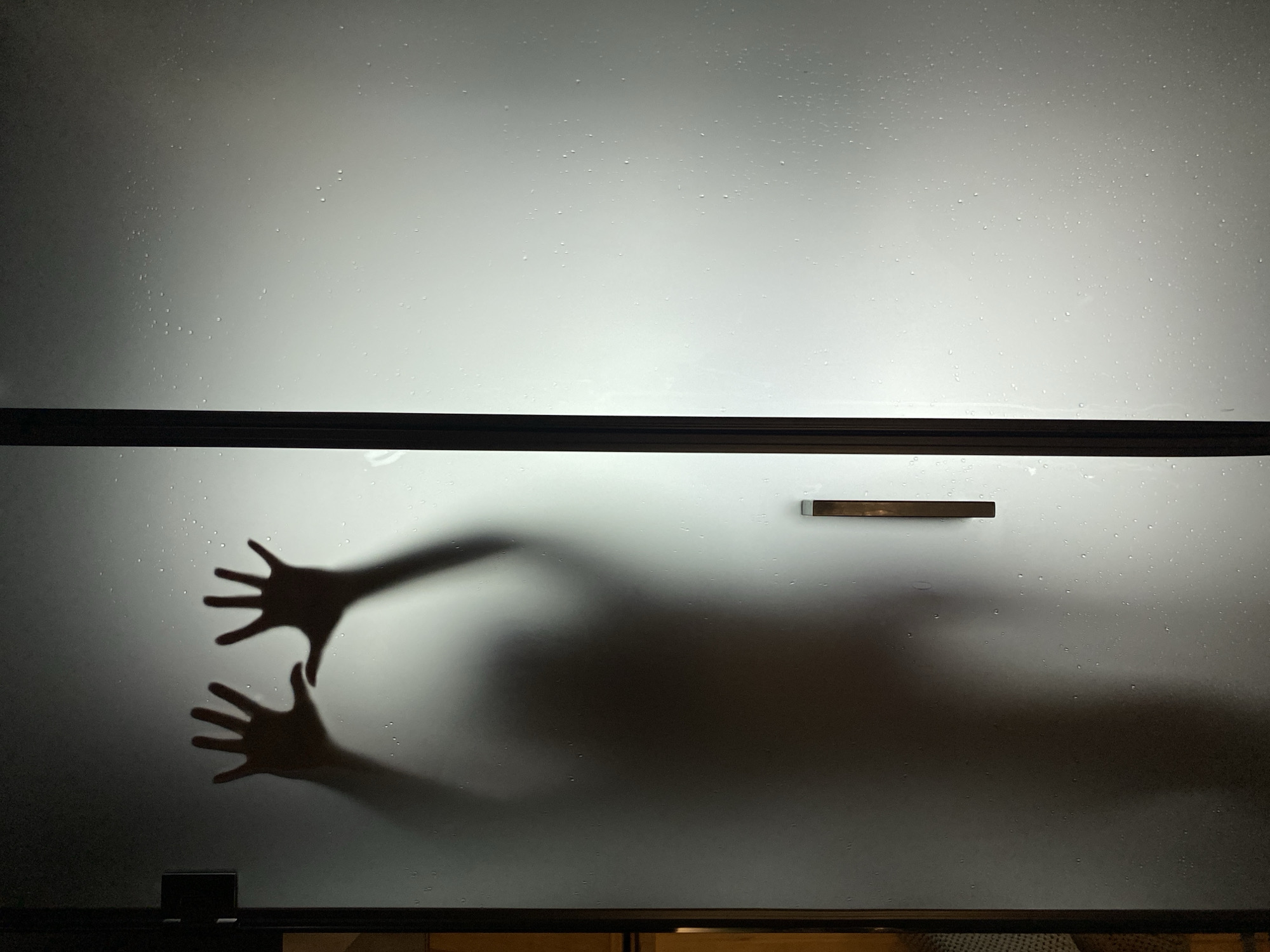 The image depicts a silhouette of a person with their hands pressed against a frosted glass surface, possibly a shower door. The silhouette is dark and indistinct, with the hands appearing as the most defined feature. Small water droplets are visible on the glass, suggesting a humid environment. The lighting is dim, casting shadows and creating a mysterious atmosphere. The image does not contain any visual elements that can be used to determine its location in Votkinsk, Russia.