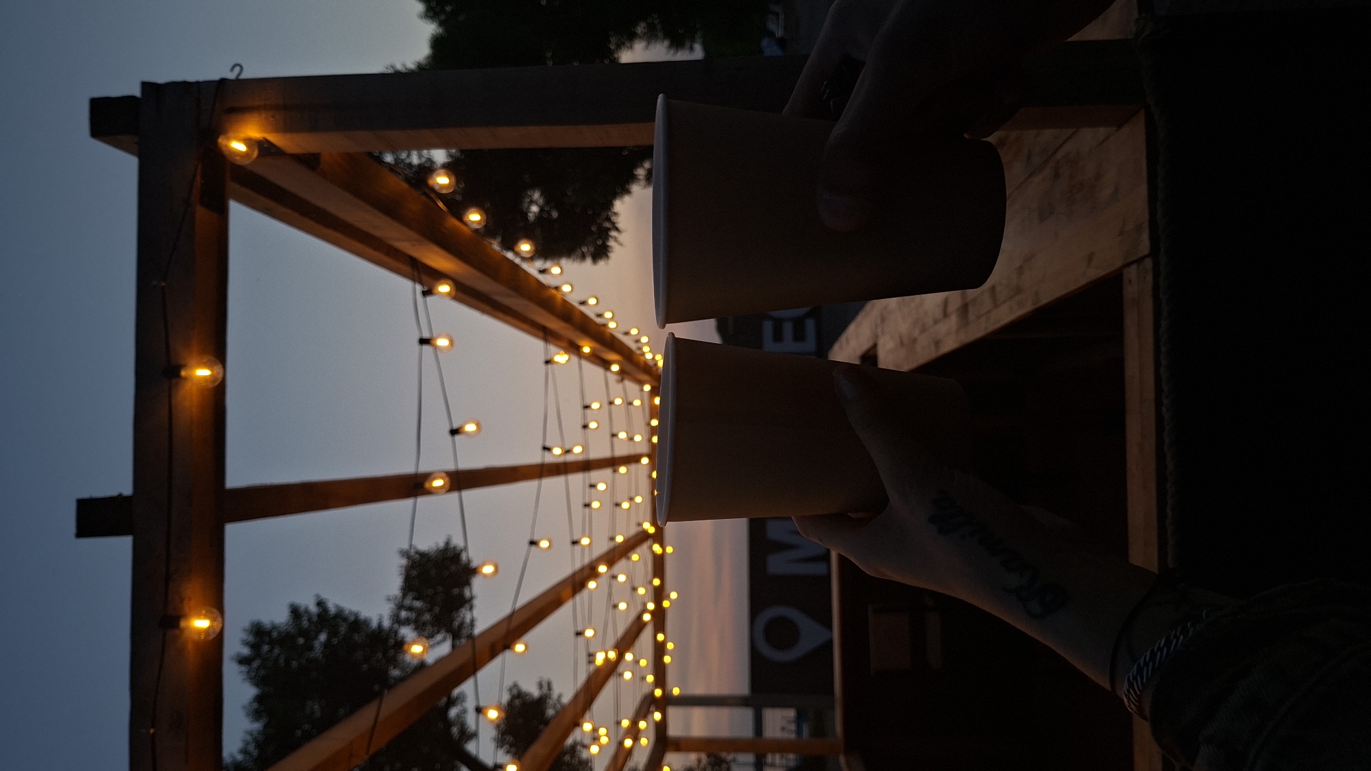The image captures a dusk scene with two hands holding disposable cups, suggesting a casual outing or event. In the background, a pergola structure is adorned with numerous string lights that are illuminated, casting a warm glow against the darkening sky. Silhouetted trees are visible on either side. The overall ambiance is relaxed and possibly hints at an outdoor cafe or gathering space. The specific location within the city of Myadzyel, Belarus, is not discernible from the visual information provided in the image.