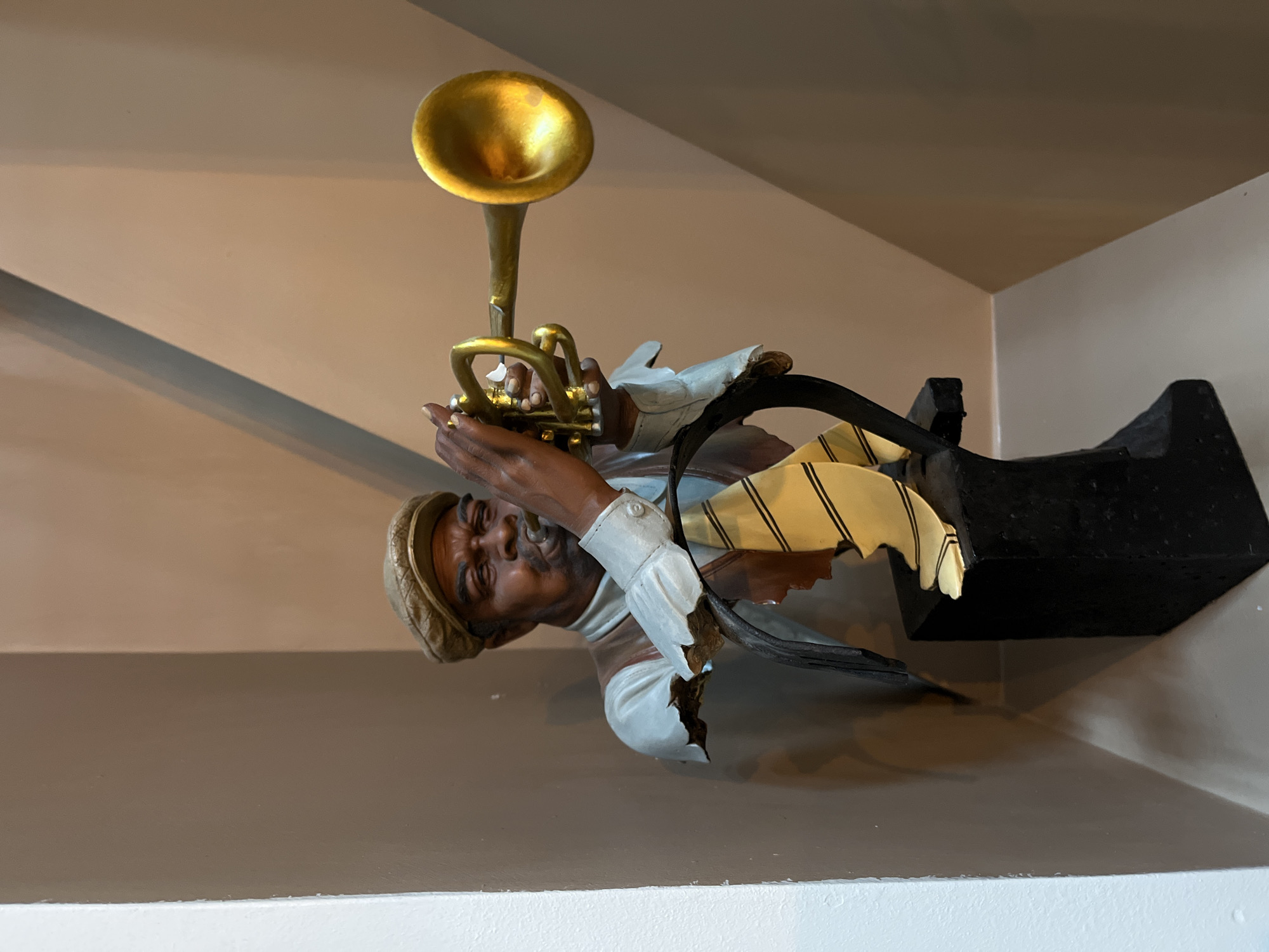 The image shows a bust of a Black man playing a golden trumpet. The bust is detailed, with the man wearing a cap, a vest, a shirt, and a tie with yellow and black stripes. The trumpet is held up to his lips, and his fingers are positioned on the valves. The bust is mounted on a black pedestal, which is placed on a surface. The background consists of two walls, one a light beige and the other a darker taupe, meeting at an angle. The lighting suggests an indoor setting, possibly a display or exhibition. No information is available to determine the location of this scene within the city of Petropavlovsk-Kamchatsky, Russia.