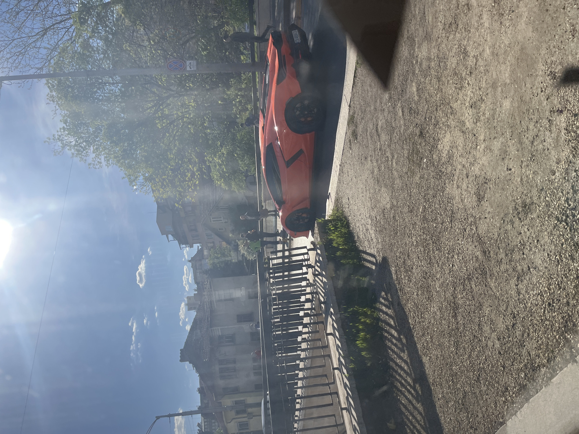 This image captures a vibrant daytime street scene in what appears to be an urban area of Bulgaria, consistent with Sofia. The sky is bright blue with scattered white clouds, and direct sunlight causes significant lens flare on the left side of the frame, creating strong shadows.
The central subject is a striking orange exotic sports car, likely a Lamborghini, parked on the side of an asphalt road. Its license plate, "PB 0086AA", is clearly visible; "PB" is a common prefix for Plovdiv, Bulgaria. Several blurred figures of people are seen near the car and further down the street, some appearing to be walking or standing casually.
To the right of the parked car, there's a gravel or dirt path alongside a dark metal fence or railing. This fence casts long, distinct shadows across the path, indicating strong sunshine. Beyond the fence, multi-story residential buildings with traditional European architecture are visible, interspersed with green trees that suggest springtime or summer. A street sign displaying a red circle with a blue diagonal line (indicating no stopping or parking) and the number "4" is affixed to a pole above the car. The scene depicts a typical sunny day in a Bulgarian city.