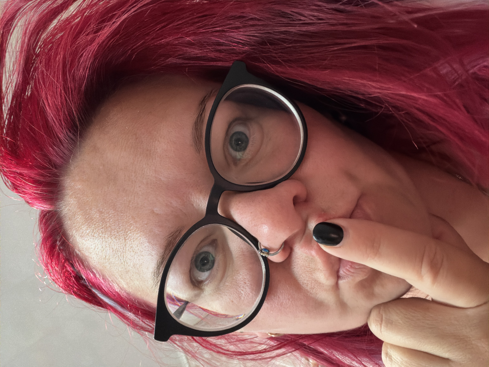A woman with bright pink hair and black-rimmed glasses is looking at the camera with her index finger pressed to her lips. She has a nose ring and is wearing dark nail polish on her right index finger. The background is a plain, light-colored wall. The image was taken indoors.