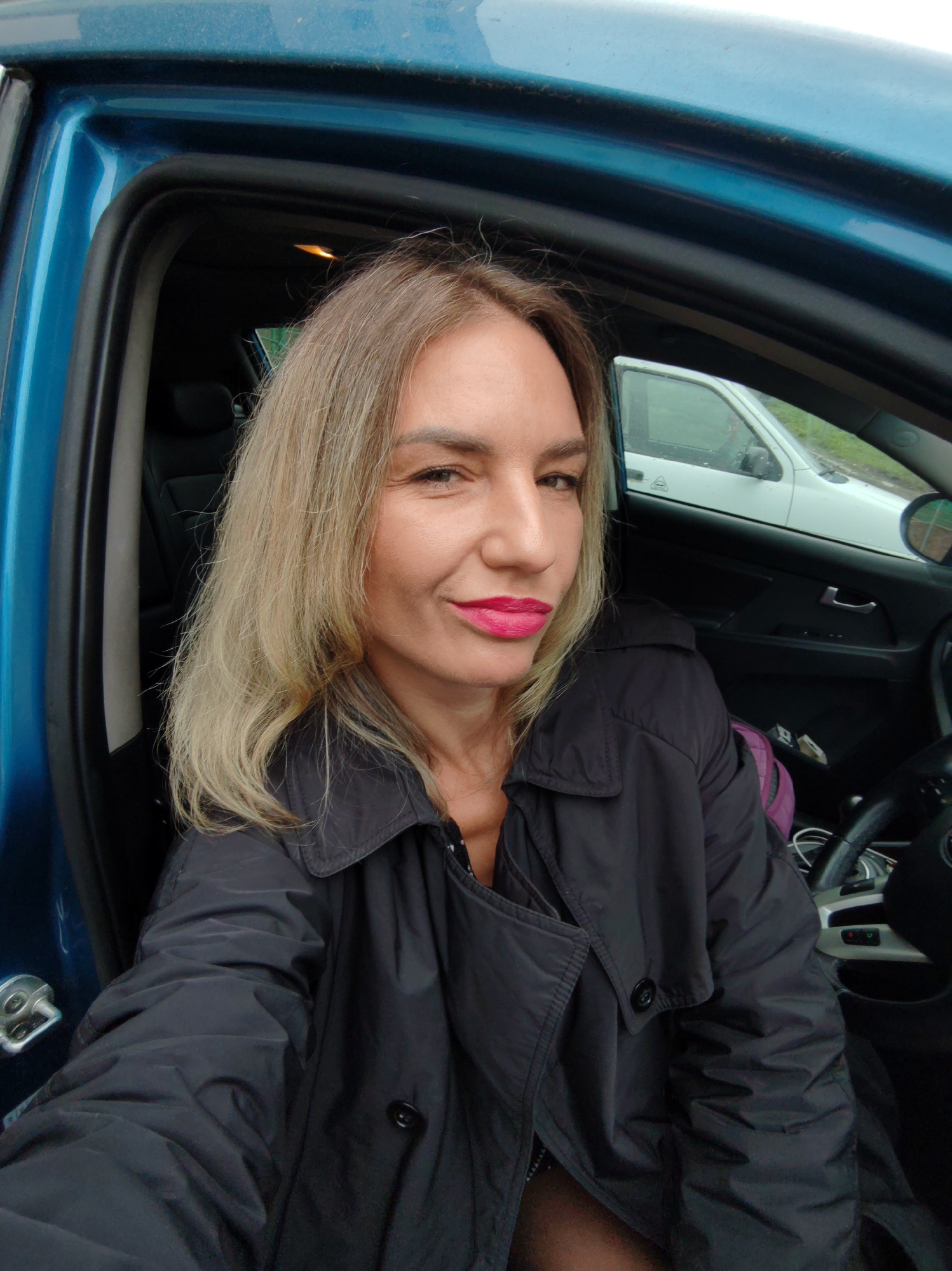 A middle-aged Caucasian woman with light blonde hair and pink lipstick is seated in the driver's seat of a blue car. She is wearing a black trench coat. The interior of the car, including the steering wheel and dashboard, is visible. Through the open car door, a white car can be seen parked outside. The location is consistent with a street setting within a city, potentially Petropavlovsk-Kamchatsky, Russia.