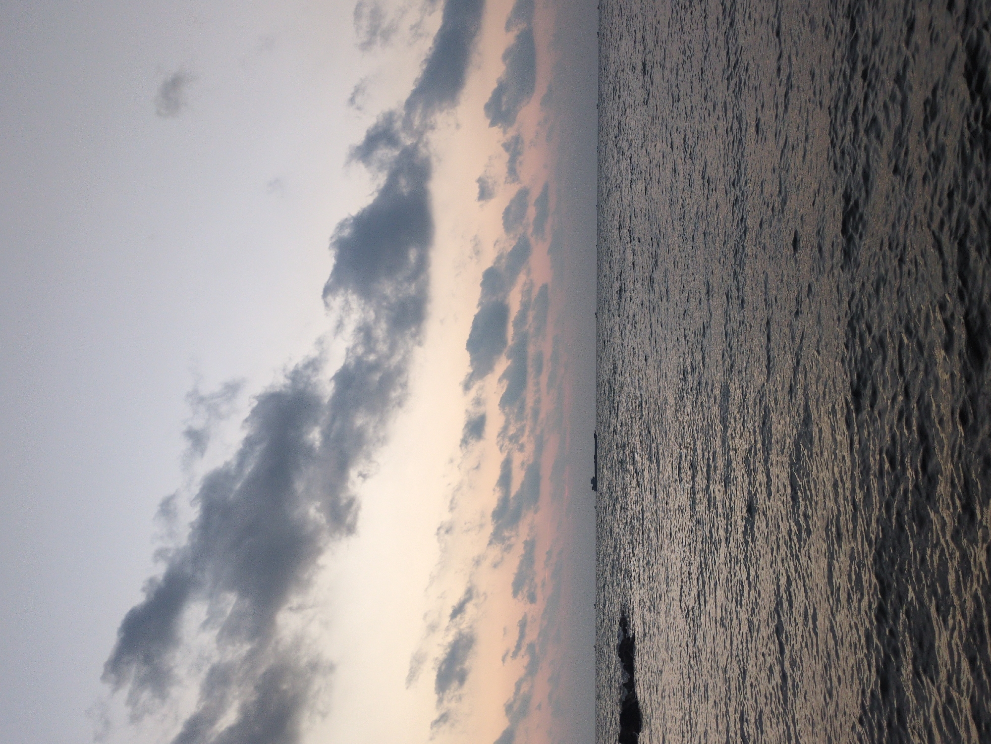 The image is of the ocean, likely in Phú Quốc, Vietnam. The left side of the frame contains clouds in the sky, colored with a mix of grays and light oranges, suggesting it might be near sunrise or sunset. The right side of the frame depicts the ocean, with textures indicating waves or ripples on the water's surface. A small ship is visible on the horizon. The image appears to be rotated, potentially taken with a mobile device. The environment suggests an outdoor setting near the coast of Phú Quốc, Vietnam.