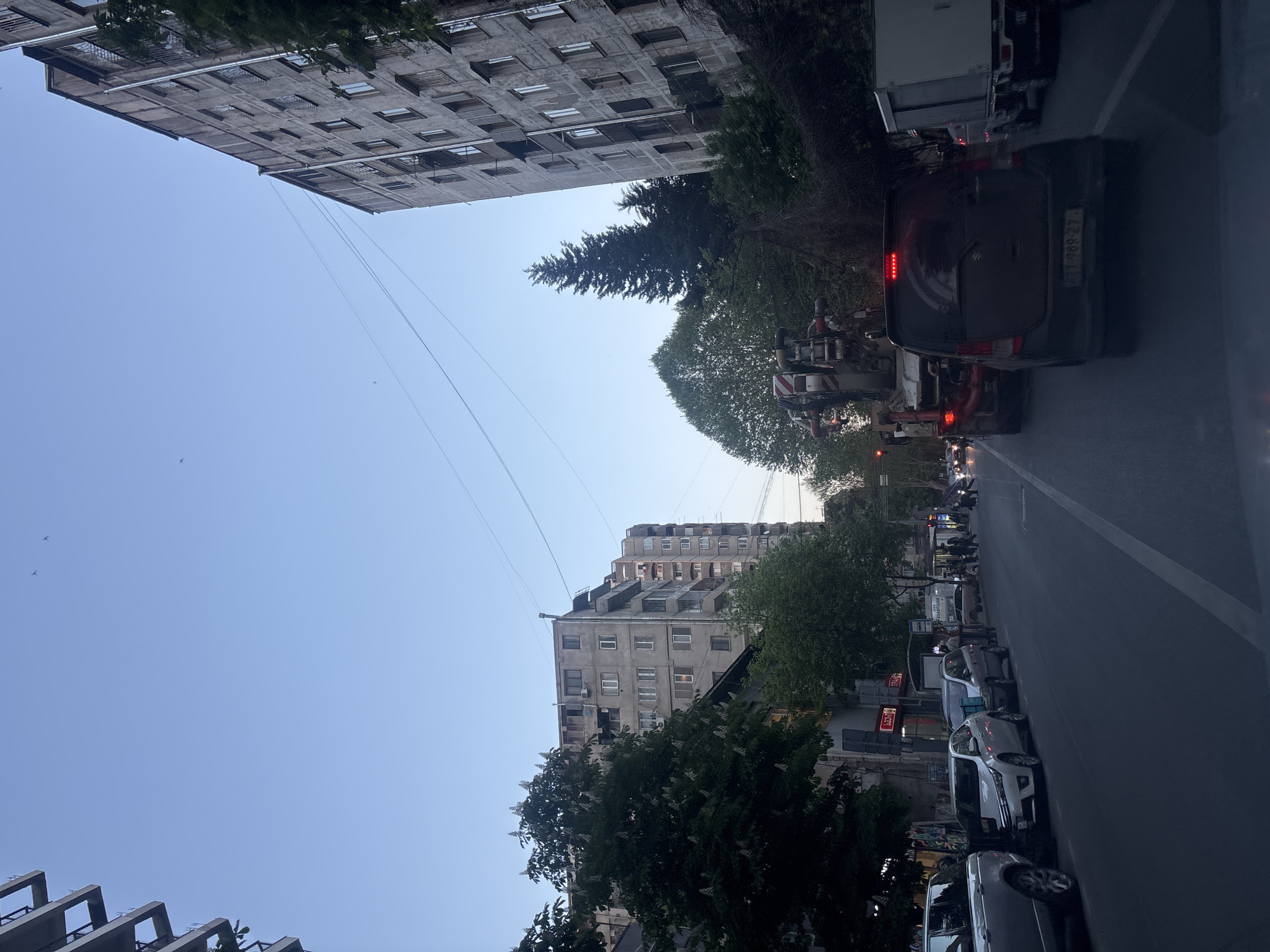 This image captures a street scene in an urban environment, viewed from a slightly elevated perspective. When correctly oriented (rotated 90 degrees clockwise), it depicts a thoroughfare lined with buildings and vehicles. The time of day appears to be late afternoon or early evening, indicated by the soft lighting, long shadows, and the clear, light blue sky.
The scene features several multi-story buildings, characteristic of city architecture, with numerous windows and balconies. Their concrete facades suggest an older, possibly Soviet-era, construction style. Green trees, including a distinct coniferous tree, are interspersed along the street and between the structures, providing natural elements to the urban landscape. Thin power or telecommunication lines crisscross the sky above.
On the street level, various vehicles are visible. Cars are parked along the right side of the road, and traffic is present in the main lane. A dark minivan or SUV is prominent, with its taillights illuminated and a license plate reading "TT-989-27." Ahead of it, a service vehicle, possibly a cherry picker or a truck with a specialized apparatus marked with red and white stripes, can be seen. Further down the street, more parked cars and a few indistinct pedestrian figures are visible on the sidewalk. Several shop signs with text, likely in Georgian, are visible on the buildings, though most are not clearly legible. One sign appears to read "PHARMA" or "CARS" in Latin script.
The overall impression is a bustling yet serene urban street in Tbilisi, Georgia, as daylight begins to fade.