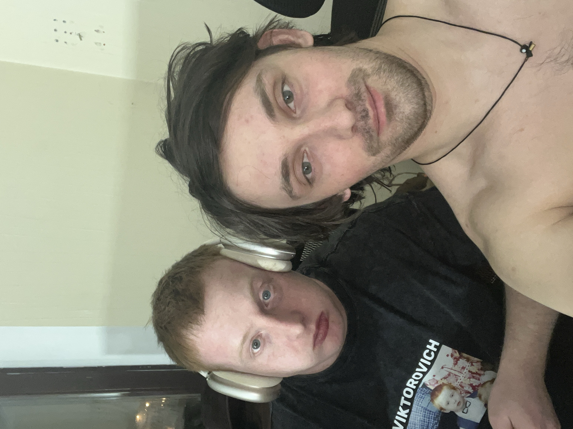 Two men are in the frame. The man at the top has long dark hair and a short beard. He is wearing a black necklace and is shirtless. The man at the bottom has red hair and is wearing headphones. He is also wearing a black t-shirt with "VIKTOROVICH" printed on it, along with a picture of a child. The background is a light-colored wall. I cannot determine the location of this image.