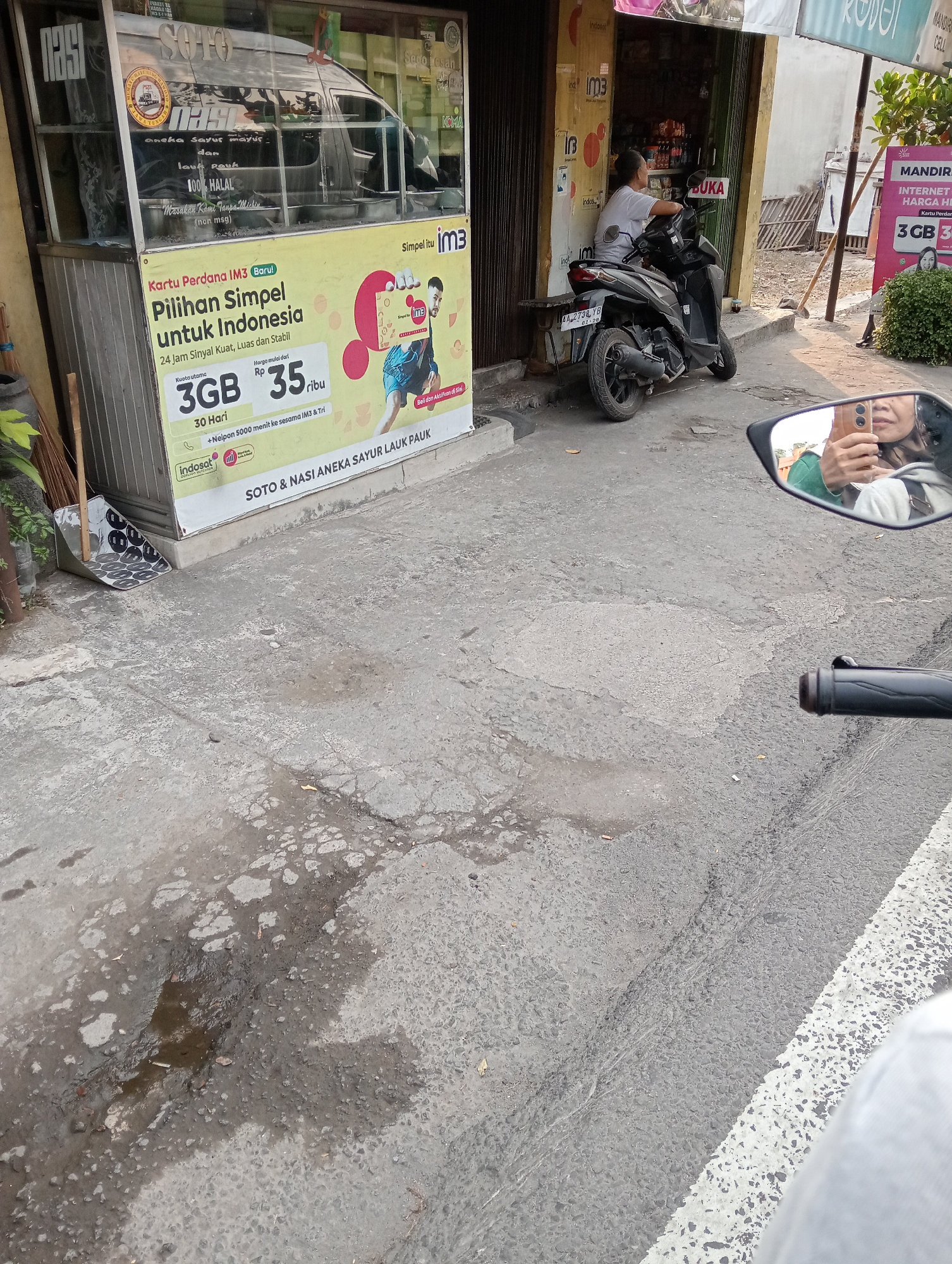 The image depicts a street scene in Salam, Indonesia, with a prominent yellow banner advertising a mobile phone plan. The banner, affixed to a food stall labeled "SOTO & NASI ANEKA SAYUR LAUK PAUK," advertises "Kartu Perdana IM3 (Baru) Pilihan Simpel untuk Indonesia" with a promotion of "3GB for 30 Hari" for Rp 35,000. Next to the banner, a black motorcycle is parked, with a person sitting on it, facing away from the camera. In the background, a store is visible with shelves stocked with products. A side view mirror of a motorcycle, with a reflection of a person holding a smartphone, is in the foreground. The street is paved with concrete, showing signs of wear and tear, including puddles and cracks. The overall environment suggests a typical street-level commercial area in an Indonesian town.
