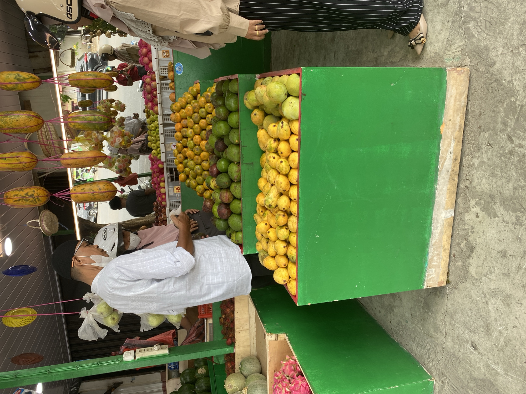 An outdoor market scene is depicted, likely in a tropical region given the produce. In the foreground, a man wearing a white patterned shirt, a black skullcap, glasses, and a face mask is interacting with a vendor. He is looking down at his hands, which are holding a piece of paper or a receipt. Next to him, a woman wearing a pink headscarf and a white helmet is partially visible. Behind them, a display of green avocados and yellow-orange mangoes is arranged in large green crates. Further back, papayas are hanging overhead. The market appears to be bustling, with other shoppers and vendors visible in the background. The ground is concrete, and the market stalls are covered by a simple roof structure.