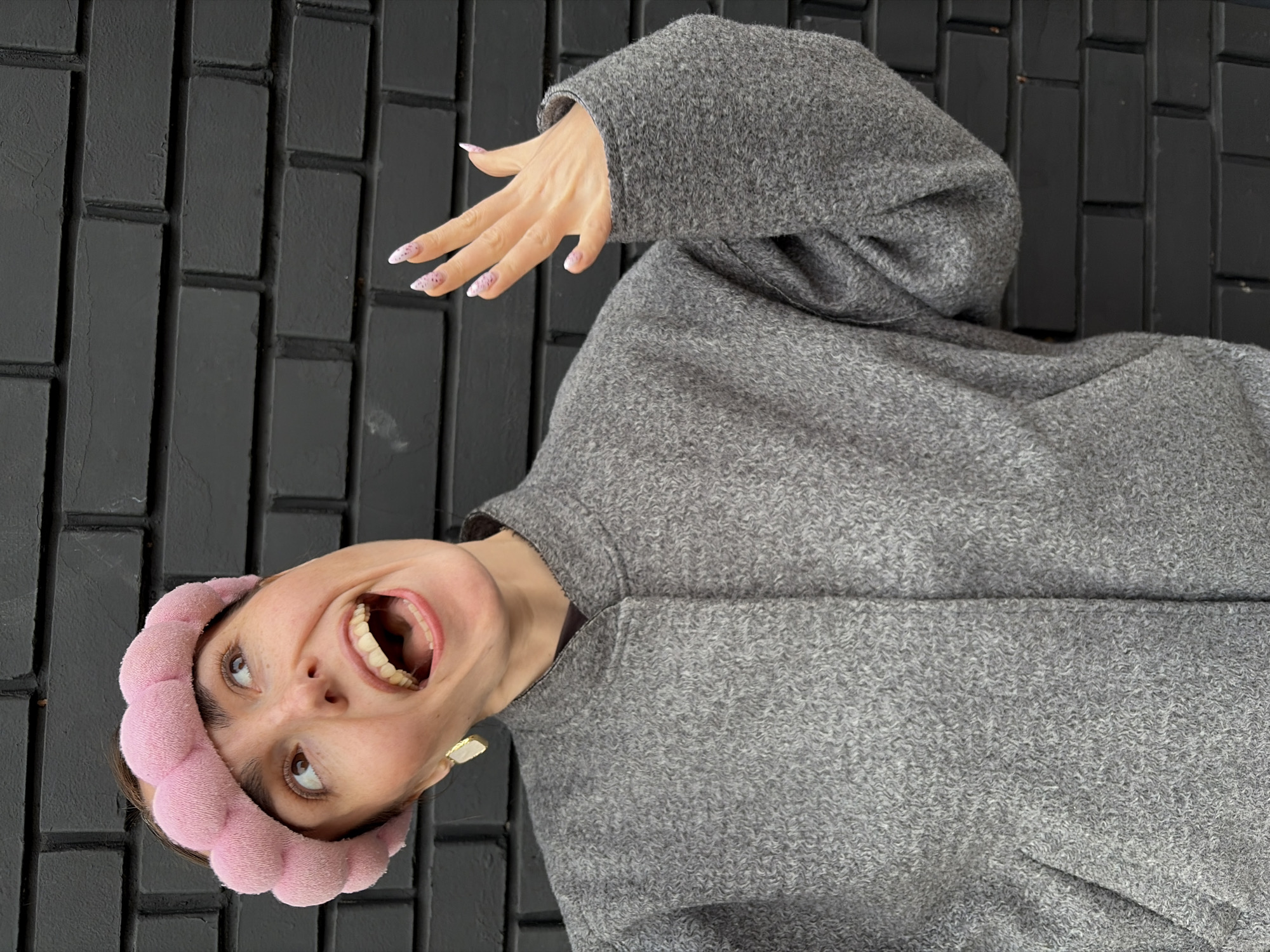 A woman with a pink, padded headband and a gray textured coat or sweater, is seen against a dark brick background. She is positioned at an angle, with her head tilted back and her mouth open in a smile or exclamation. Her hand, with manicured nails, is raised near her head. The image is not a location in Izhevsk, Russia.