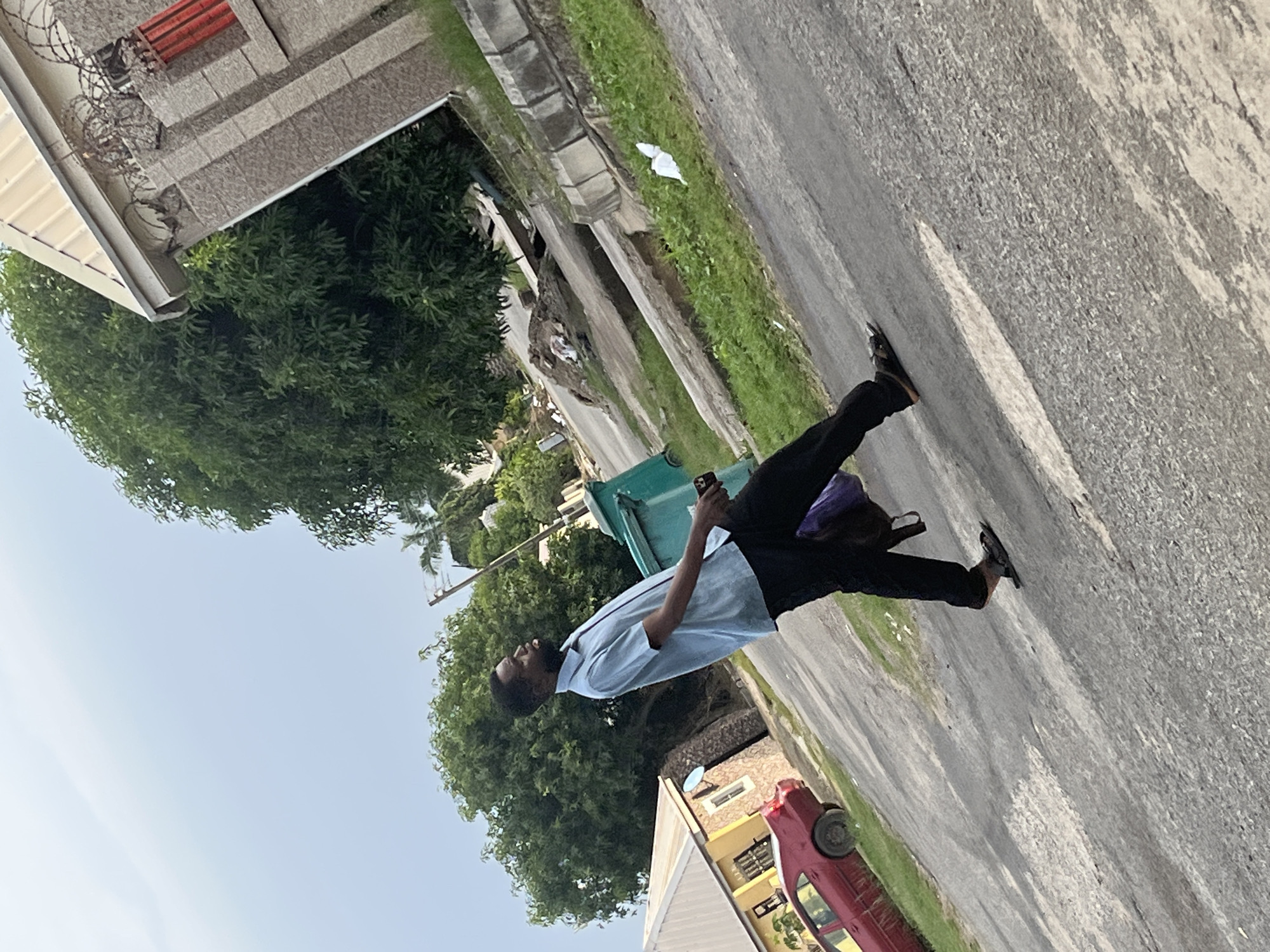The image shows a man wearing a blue shirt and black pants walking on a paved road in what appears to be a residential area in Abuja, Nigeria. He is carrying a purple bag over his shoulder and holding a dark object, possibly a phone, in his right hand. The road has a slight incline, and the surrounding area includes houses, trees, and a green garbage bin. A red car is parked on the left side of the road. The sky is overcast.