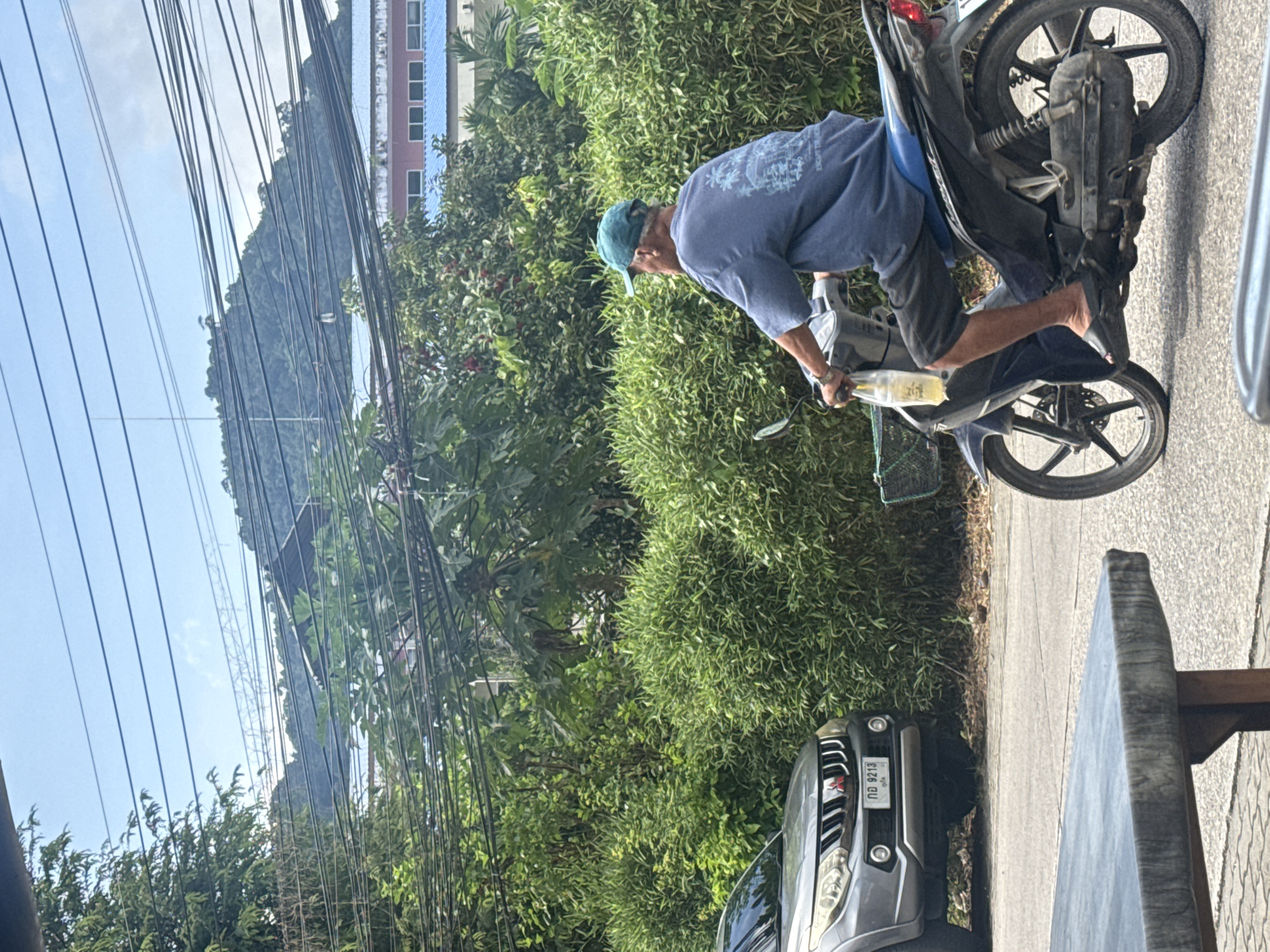 A man wearing a blue t-shirt and a teal baseball cap rides a dark-colored motorcycle in Kathu, Thailand. He is holding a bottle of yellow liquid in his left hand. To his left, a gray SUV is parked. The motorcycle is positioned on a road adjacent to a dense hedge. In the background, a hill covered in lush green foliage rises, with a building featuring a blue roof visible behind the trees. The scene is illuminated by daylight, and numerous power lines crisscross the sky. A gray table with a wooden base is visible in the foreground, partially obstructing the view of the road.