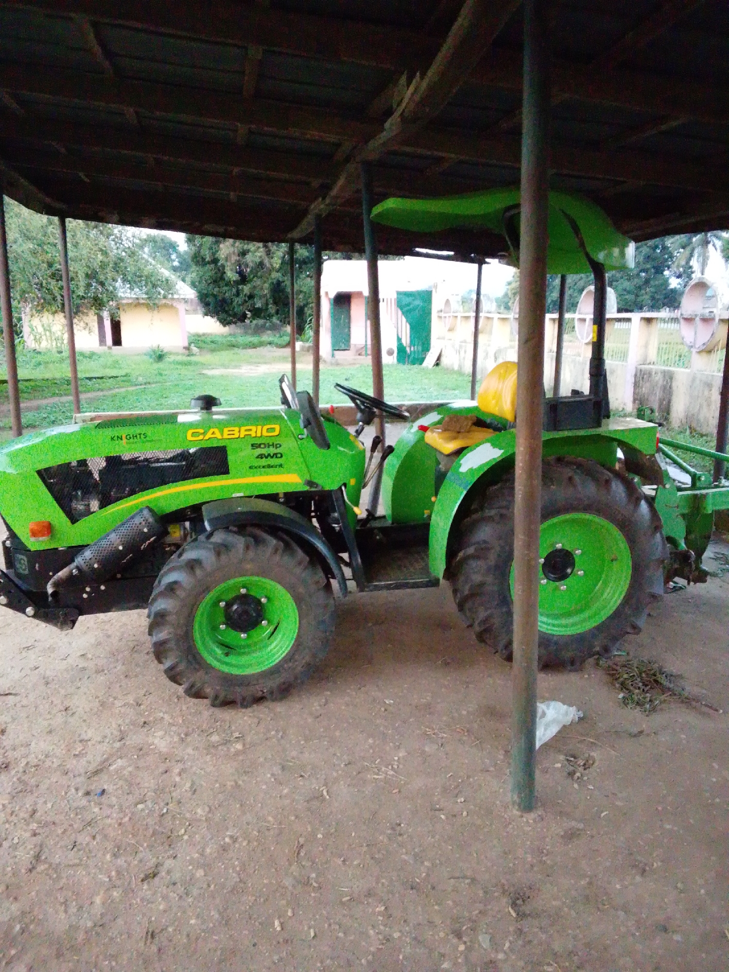 The image shows a green tractor parked under a covered structure. The tractor has the brand name "CABRIO" and "50HP 4WD" written on its side. The tractor is large and appears to be in good condition. The covered structure is made of wood and metal poles. In the background, there are some buildings and trees. The ground is covered with dirt and some scattered debris. The scene appears to be in a rural or semi-rural area. The provided location of Serti, Nigeria is plausible given the visual cues. There are no people or animals visible in the image. The overall atmosphere is one of stillness and inactivity.
