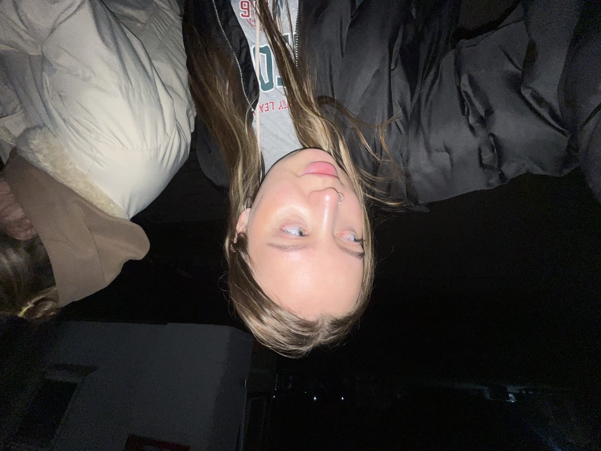 The image shows a person with light skin and light brown hair wearing a gray shirt with a graphic design, a black jacket, and a nose ring. They are in an upside-down orientation. The background shows an off-white, puffy object (possibly a coat or blanket) to the left, and a building and dark sky in the distance. The location, Novopokrovskaya, Russia, is not confirmed by the image content.
