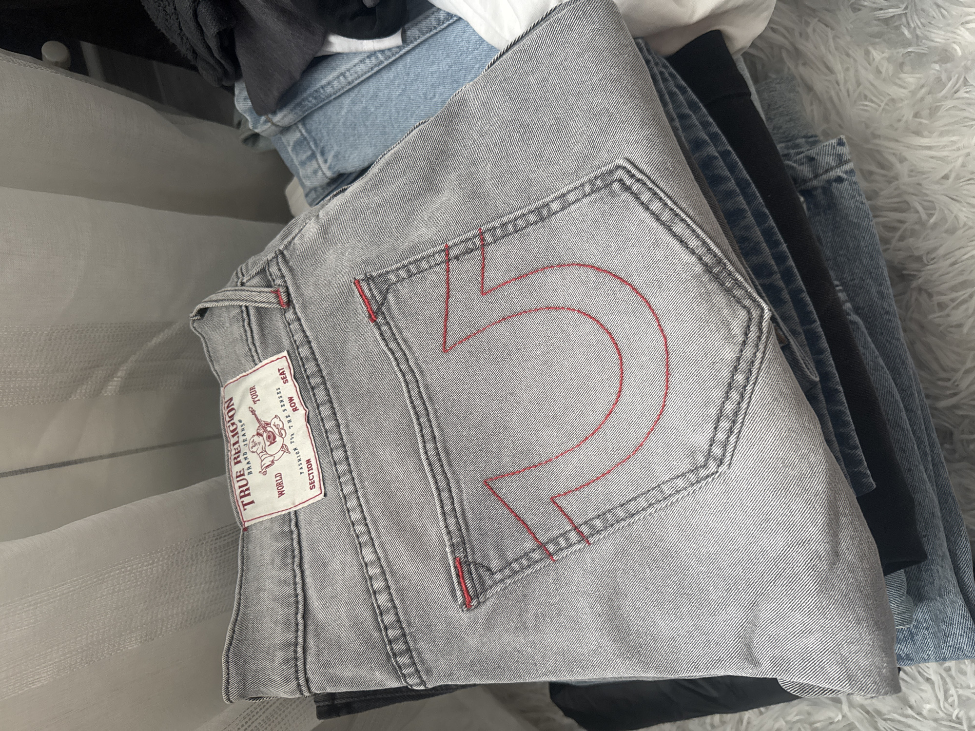 The image displays a close-up, overhead view of a folded pair of light grey True Religion denim jeans, prominently featuring its distinctive back pocket and branding. The jeans are made of a light grey denim with a visible diagonal twill weave. The back right pocket is adorned with bold red stitching, forming a stylized horseshoe or "U" shape. Double grey stitching outlines the pocket and runs along the seams of the jeans.
A red and white brand patch is visible on the waistband of the jeans, located above the back pocket. Text on the patch includes "TRUE RELIGION", "BRAND JEANS®", "WORLD", "TOUR", "FASHION FOR THE SENSES", "ROW", "SEAT", and "SECTION".
The jeans are stacked or folded among other clothing items. To the right and behind the grey jeans, other denim garments, including light blue jeans and a black fabric item, are partially visible. A soft, fluffy white texture, possibly a blanket or rug, appears in the background on the right. On the left side of the frame, a translucent white fabric with faint horizontal stripes, possibly a curtain or part of a laundry basket/container, is present. The overall setting appears to be indoors, likely in a bedroom or laundry area, with no specific time of day or weather discernible. No people are visible, and no activity is occurring; the image captures a static arrangement of garments.
