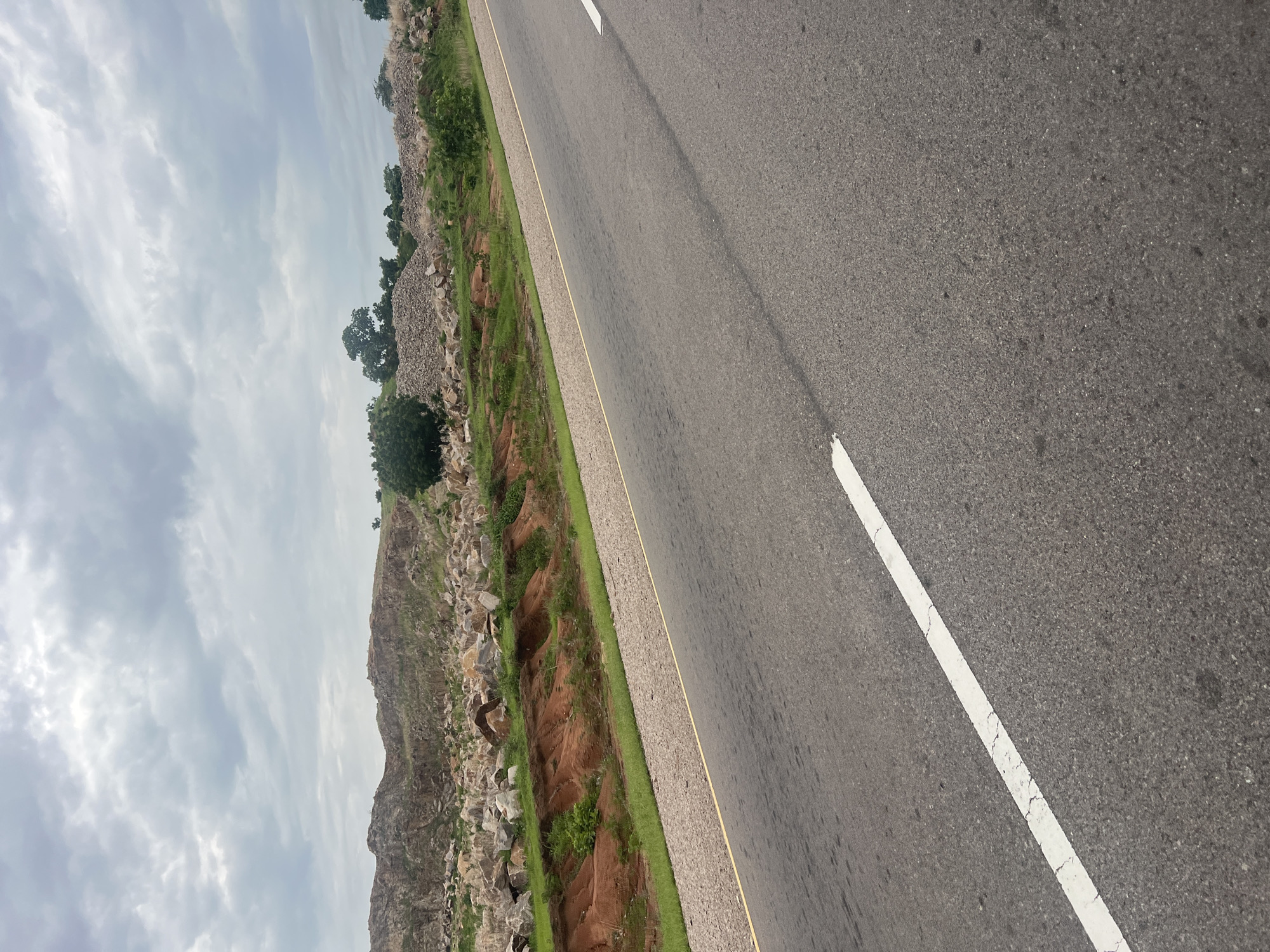This image shows a paved road with a white line marking the lane. The road is flanked by a grassy verge and a rocky, uneven terrain. In the distance, there are trees and the sky is overcast with clouds. The image is taken from a slightly elevated perspective, looking down the road. The time of day appears to be daytime, and the weather is cloudy. The location is Minda, Nigeria. There is no visible text in the image. The scene depicts a rural or semi-rural setting, likely a highway or a major road connecting different areas in Nigeria. The rough terrain and vegetation suggest a natural landscape.