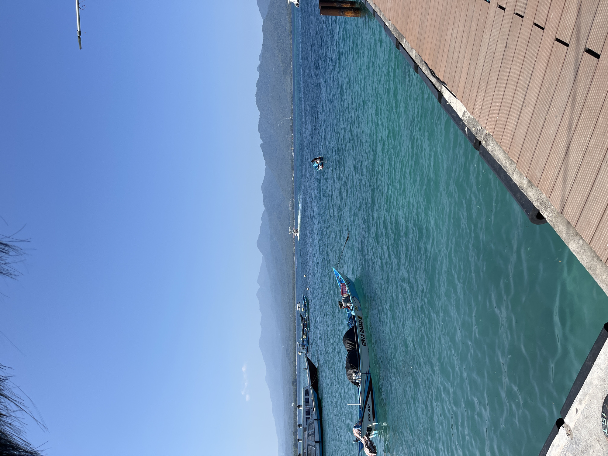The image shows a view from a wooden dock overlooking a body of water, likely an ocean or large bay, in an area of Unknown, Indonesia.
The water is a vibrant turquoise and slightly disturbed, suggesting a gentle breeze or current. Several boats are docked parallel to the dock, with visible names, including "CINTA DAMAI 3" and "GILI AIR" on at least two of them. Some of the boats appear to be traditional outrigger canoes.
In the distance, a mountainous coastline is visible under a clear, blue sky. A single person is seen swimming in the water between the dock and the coastline.
The dock is made of wooden planks, and the perspective suggests a relatively high vantage point. The overall scene suggests a tropical or coastal environment, likely a tourist destination or harbor.