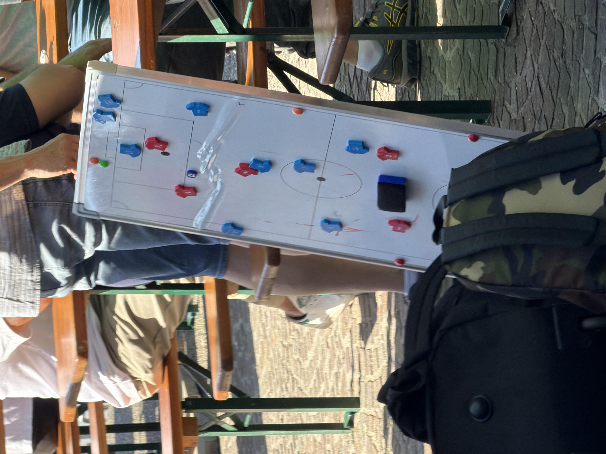 The image depicts a whiteboard set up on a table at an outdoor seating area, likely at a restaurant or cafe. The whiteboard displays a diagram representing a soccer or hockey game formation, with blue and red magnetic pieces arranged to represent players. A black eraser is also visible on the board. Several individuals are seated around the table, indicated by their visible clothing, including denim shorts, khaki pants, and a white shirt. A backpack, partially obscured in the foreground, has a camouflage print on its upper portion and is black on the lower portion. The location of the scene is Fentange, Luxembourg.
