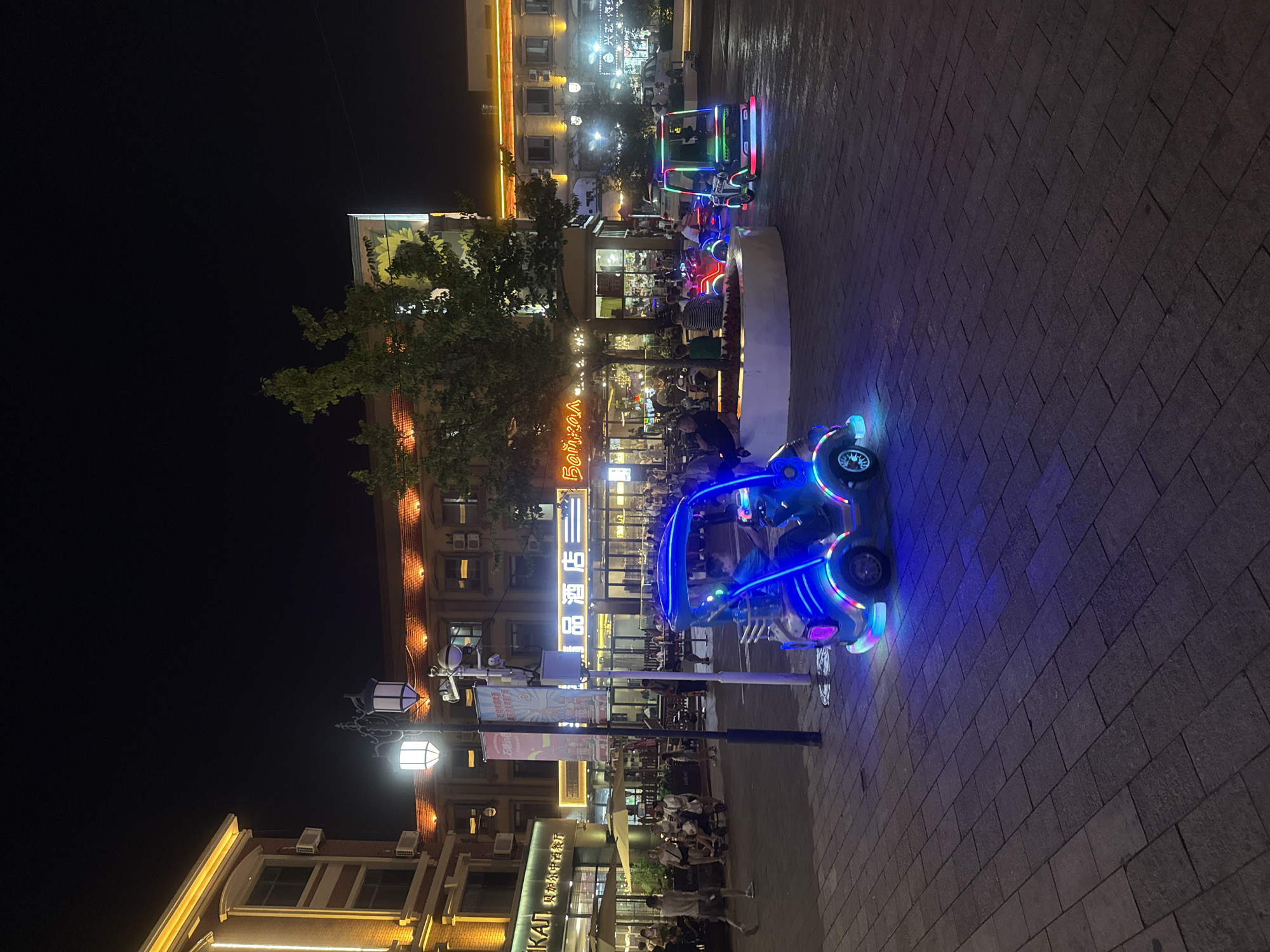 This image depicts an outdoor pedestrian area at night, likely in a commercial district of Qinhuangdao, China, given the presence of signage with Chinese characters. The scene features several illuminated ride-on toys for children, resembling small cars, scattered across a paved walkway. One prominent blue toy car in the foreground is brightly lit with neon blue and purple lights. Another toy car, green and red lit, is visible in the middle ground. In the background, buildings line the walkway, displaying various illuminated signs and architectural lighting, suggesting they house businesses such as restaurants and hotels. Several people are present, some walking and others seated at tables outside the establishments. The overall atmosphere is lively and illuminated, typical of an evening entertainment or shopping area.