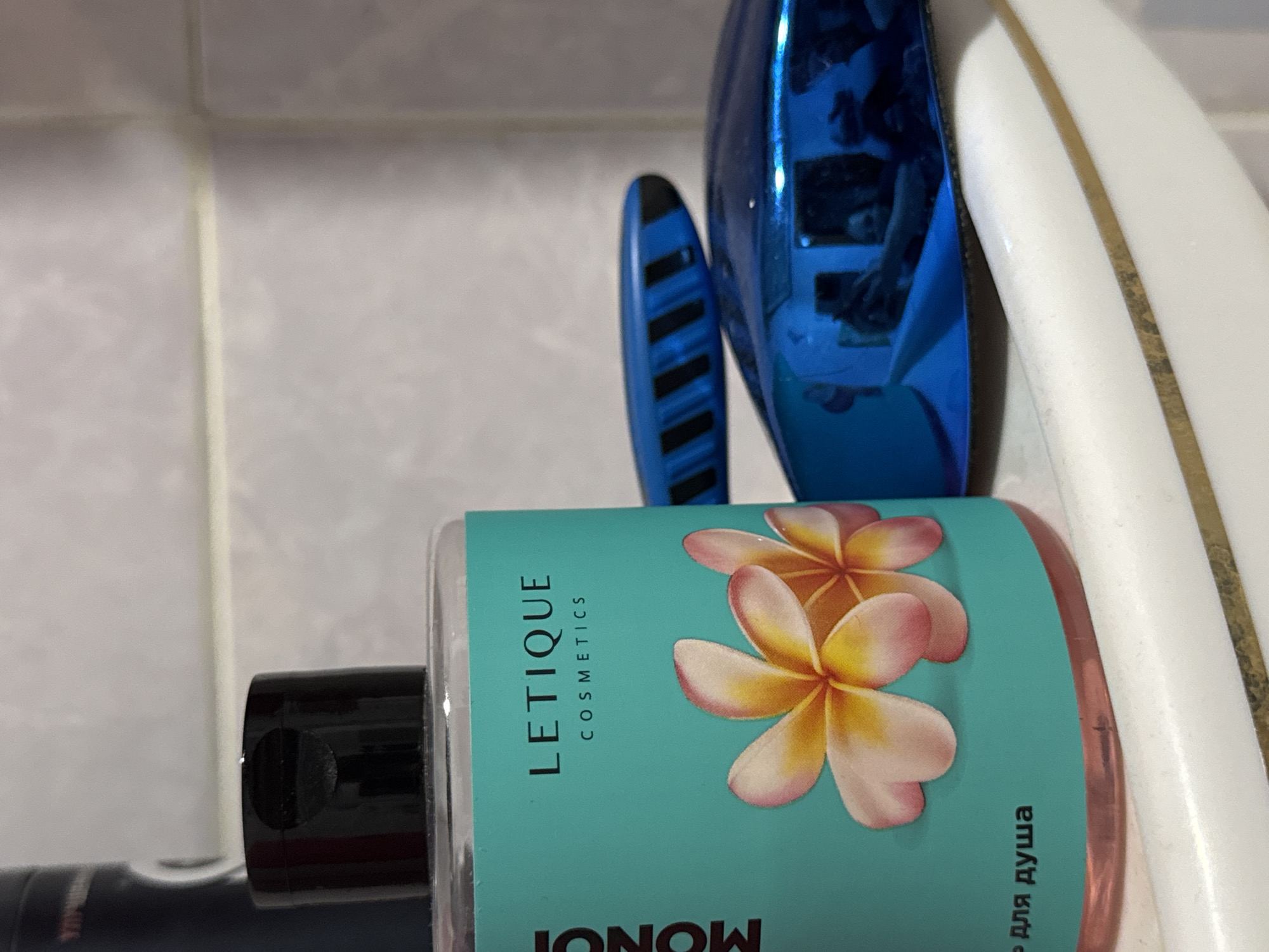 The image shows a close-up of cosmetic products on a bathroom shelf. The primary subject is a clear plastic bottle with a turquoise label featuring pink and yellow plumeria flowers. The brand name "LETIQUE COSMETICS" is visible on the label, along with the word "MONOI" vertically oriented on the left side. Below the flowers, the Russian text "Р ДЛЯ ДУША" which translates to "for shower" is present. To the right of the bottle, two blue objects with black striped patterns are visible. The background consists of a tiled wall, likely in a bathroom. The scene is static and does not depict any specific action or interaction between people. The lighting is moderate, highlighting the glossy surfaces of the products. The location in Яромінскі сельскі Савет, Belarus is not evident from the visual information in the image.