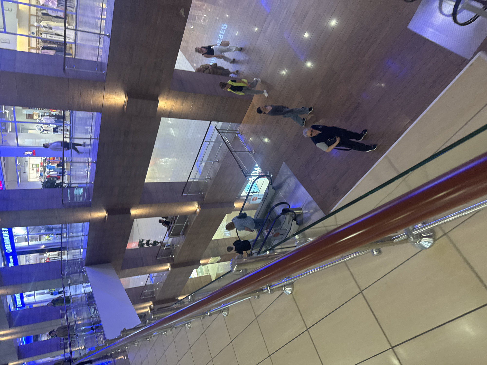 The image shows a multi-level indoor shopping mall, likely in Saint Petersburg, Russia. The architecture features modern design with wooden flooring, glass railings, and blue-toned lighting. Several people are visible on different levels, engaged in various activities.
On an upper level, several individuals are walking. One person wears a bright yellow vest, another a light-colored top and dark pants, and a third is in dark clothing. Further up, two people are seen walking, and one appears to be carrying a bag. On another floor, a person in dark clothes with gray hair is holding a bag and looking down.
A central feature of the mall is an escalator. Two people are riding the escalator, one in a blue shirt and the other in dark clothing. In the background, there are storefronts displaying clothing on mannequins. Some of the storefronts have visible text. On the left side of the image, a blue sign with white text that appears to be Russian can be seen on a store. Another store on the left has the word "BEBE" visible in large white letters.
The lighting suggests it is daytime, but the indoor setting makes the time of day less definitively indicated. The overall atmosphere appears to be a typical day at a shopping center with people browsing and moving between levels.