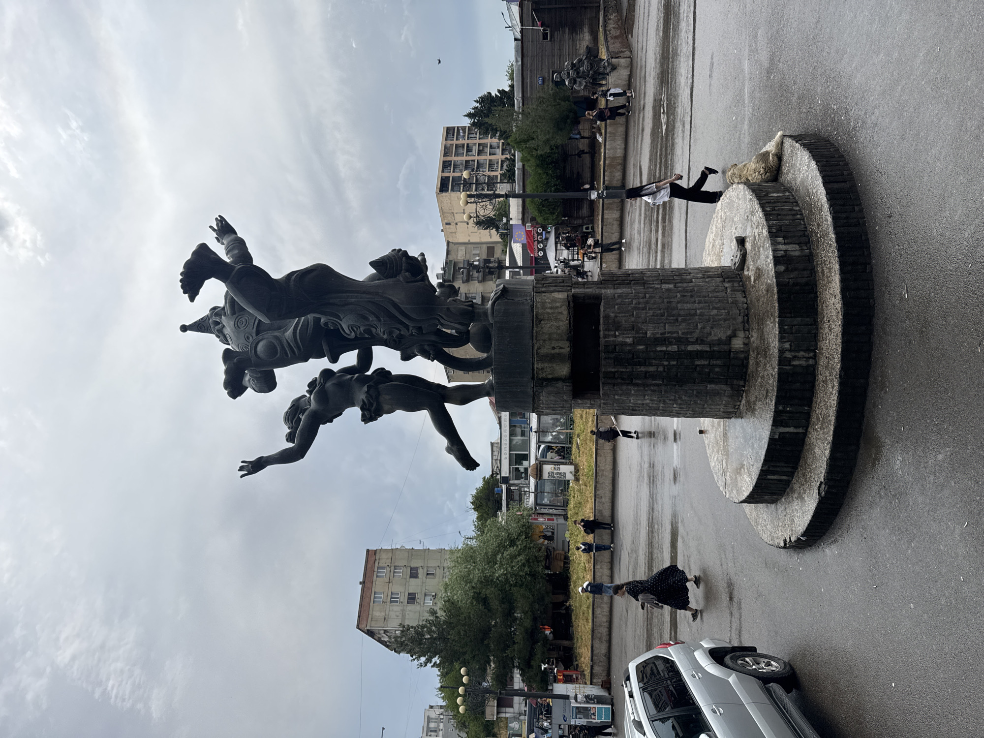 In the city of Tbilisi, Georgia, a large bronze sculpture of figures in dynamic poses dominates the scene, standing on a tiered concrete base. The sky is overcast and the ground is wet, suggesting recent rain. A silver SUV is parked to the left of the frame. Several pedestrians are visible in the background and midground, some walking, others standing. Among them, a woman in a black dress with white polka dots walks across the wet pavement. Another woman in a white shirt and black pants walks by the base of the sculpture, a fluffy light-colored dog lying near her. Buildings, some appearing to be apartment blocks, and trees are visible in the background, along with streetlights and what look like bus stops. The overall atmosphere is muted due to the cloudy weather.