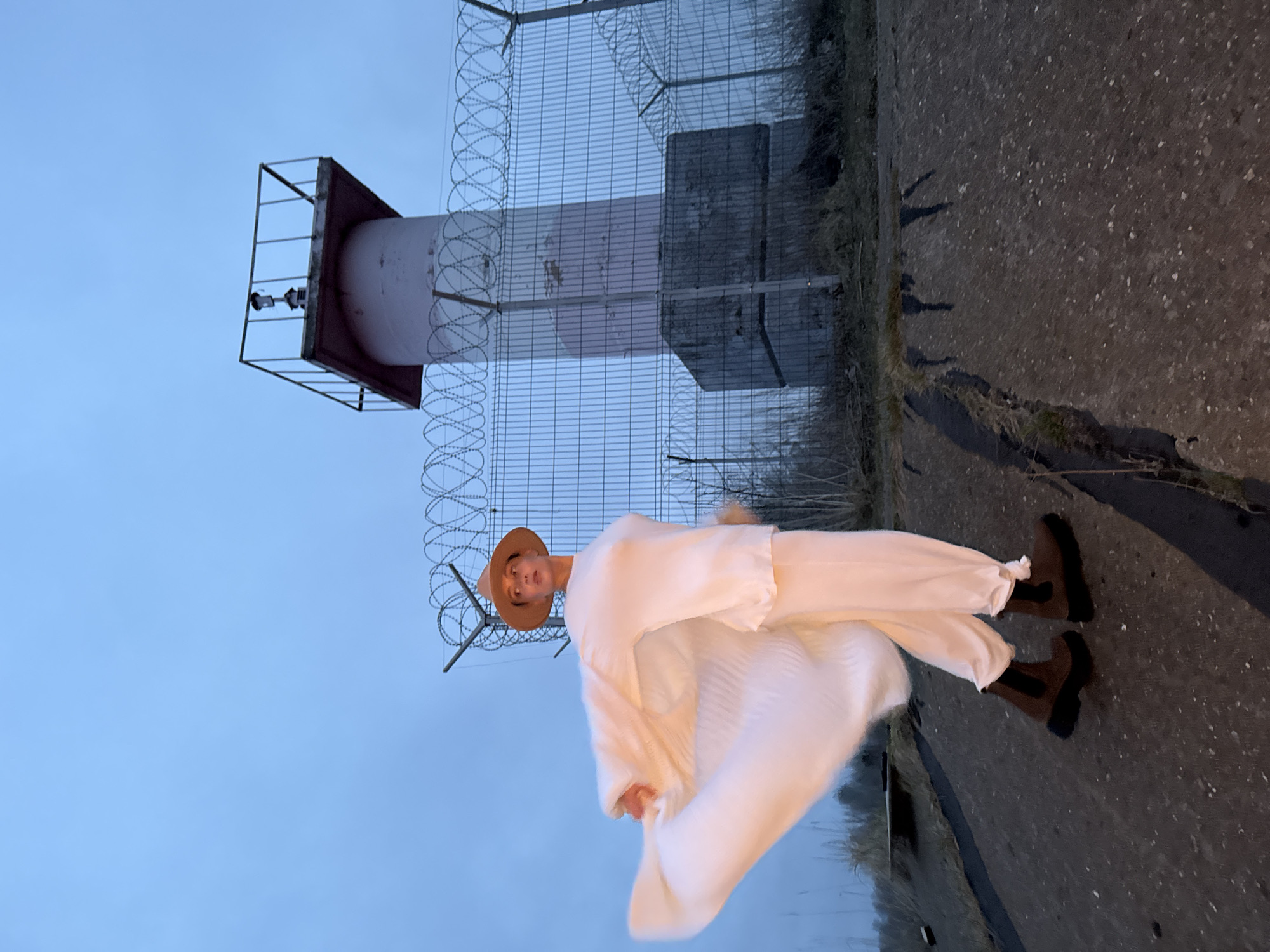 A full-length outdoor portrait captures a person standing next to a tall, cylindrical structure, likely in Chaikovsky, Russia. The individual, positioned slightly right of the frame, wears a flowing, oversized light-colored (appearing white or cream) top and matching wide-legged pants, along with a light brown wide-brimmed hat and dark brown boots. They are looking towards the camera with a calm expression, posing for the photograph.
The background is dominated by a pale-colored tower, possibly an industrial or observation tower, featuring a square metal platform at its summit equipped with what appear to be surveillance cameras or sensors. A tall wire mesh fence, topped with coiled barbed wire, runs prominently alongside the tower, extending across the scene and suggesting a restricted or secured area. The ground is a dark, textured surface, possibly concrete or asphalt, with sparse, dry vegetation along its edges.
The sky is a uniform, pale blue, indicating diffused lighting characteristic of early morning, late afternoon, or an overcast day. The setting suggests a functional or industrial environment. No discernible text is visible in the image.