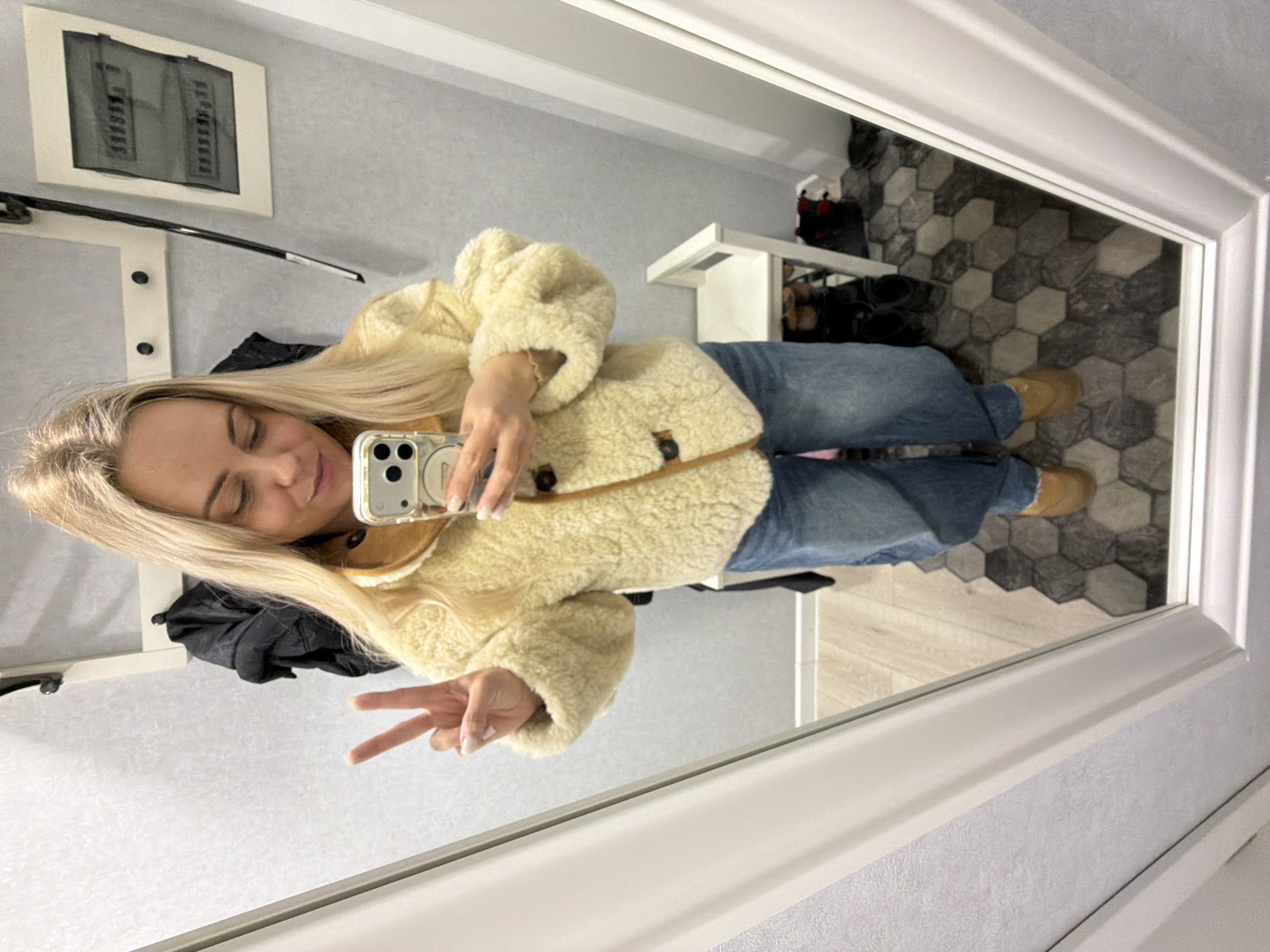 A selfie taken in a mirror reveals a light-skinned woman with long, blonde hair, wearing a cream-colored, shearling-style jacket and blue jeans. She holds a smartphone in her right hand, using it to capture the image while making a peace sign with her left hand. The mirror's reflection shows an interior space with a gray and white hexagonal tiled floor, a white table, and a coat rack with a black coat. A framed picture hangs on the wall. The specific location in Votkinsk, Russia, cannot be determined from the available information.