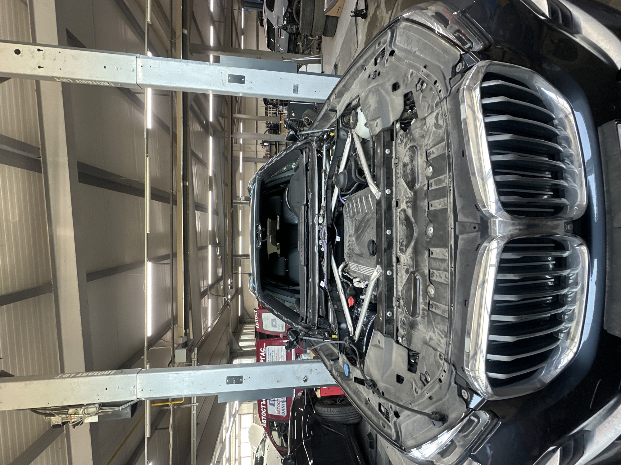 The image captures a black BMW car undergoing maintenance or repair within a professional garage or service center. The car's front end is significantly disassembled; its hood is open, and the front bumper and parts of the fascia have been removed, revealing the engine bay components including the engine cover with the "BMW" emblem, various fluid reservoirs, wiring harnesses, and structural elements. A prominent chrome-rimmed kidney grille is visible on the passenger side of the car's front.
The setting is an indoor industrial space featuring a high ceiling equipped with fluorescent lights and corrugated metal walls. A robust car lift, identified by the partially visible text "ROTARY LIFT" on its main beam, spans the scene overhead, indicating specialized automotive equipment. In the background, red workshop cabinets or equipment can be seen, bearing fragmented text such as "АВТОСТ" (likely part of 'Автосервис' - car service) and "DANIEL". No individuals are present in the frame. The scene suggests a paused or ongoing repair operation on the vehicle's front section. The image is oriented vertically, providing an aerial view of the car and the workshop environment.