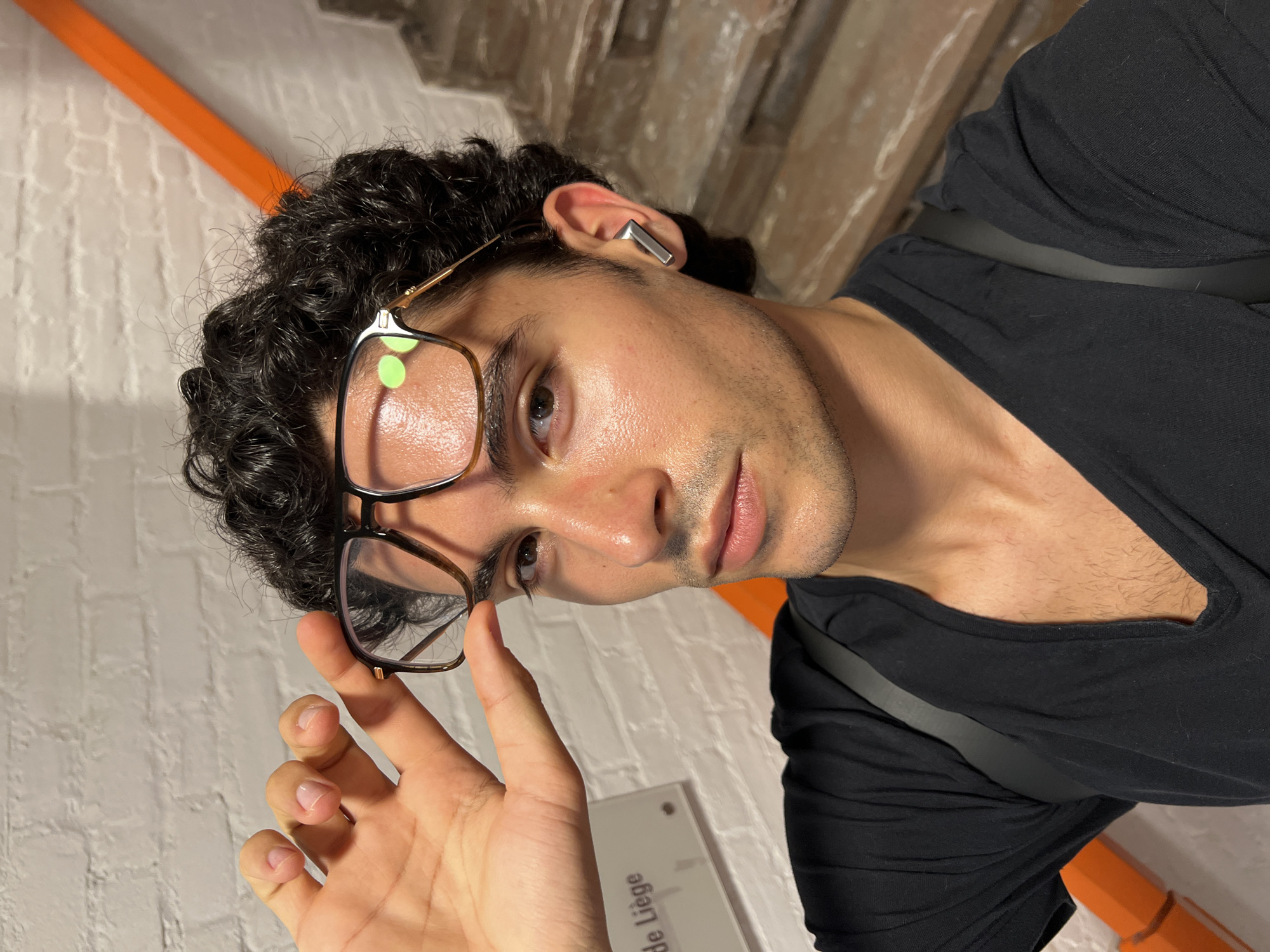 A close-up selfie shows a young man with dark, curly hair and glasses. He is wearing a black t-shirt. He is holding his glasses up with his left hand, revealing his forehead and eyebrows. His right ear has a silver earbud. In the background, there is a white brick wall and a glimpse of an orange railing and stairs. A sign on the wall reads "de Liège," indicating the location is in the city of Liège, Belgium. The lighting suggests an indoor setting, possibly a staircase or hallway.
