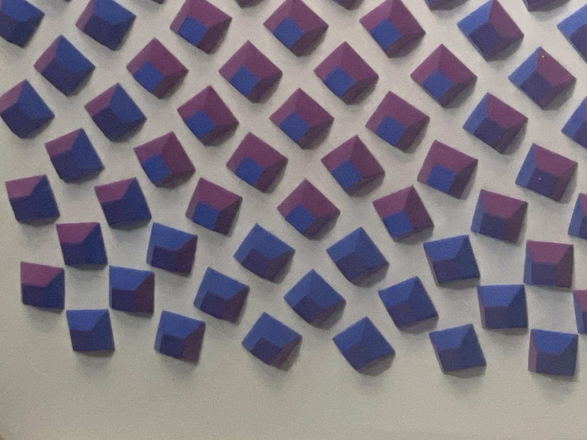 The image displays a pattern of small, three-dimensional geometric shapes, likely cubes, arranged in a grid-like fashion on a white surface. The cubes are oriented at an angle, presenting two visible faces. These faces are colored in a pattern of alternating blues and purples. The arrangement shows a gradual shift in color dominance from left to right and top to bottom. The cubes are not perfectly aligned, adding a degree of irregularity to the pattern. There is no clear indication of the location in the city of Caracas, Venezuela or any people/subjects/actions.