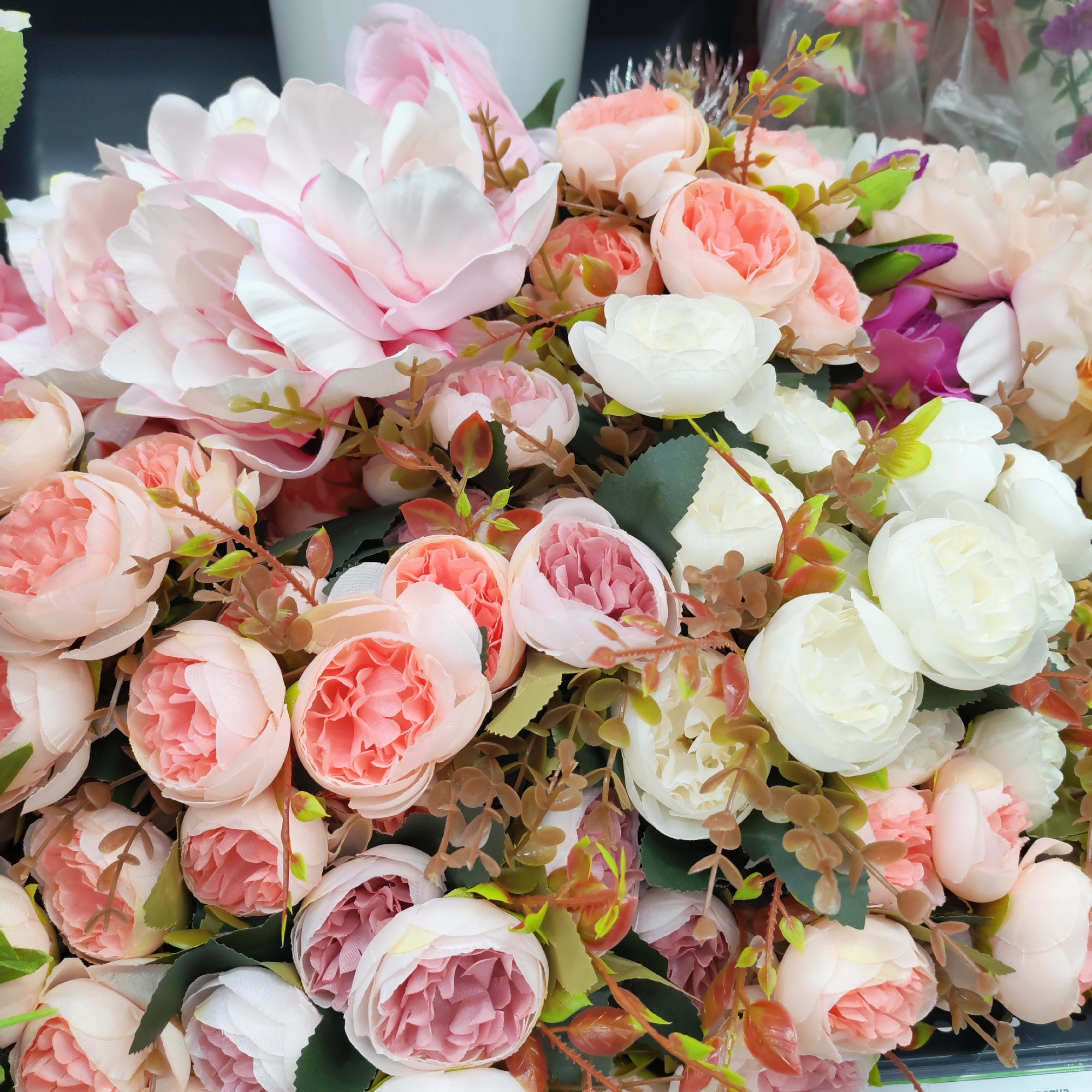The image shows a close-up of a bouquet of artificial flowers. The flowers are predominantly pink and white, with varying shades and textures. The arrangement includes larger peony-like blossoms and smaller, rose-like buds. Green and brown artificial foliage fills the spaces between the blooms. The composition is dense, with flowers overlapping and filling the frame. The specific location in Chaikovsky, Russia is not discernable from the image.