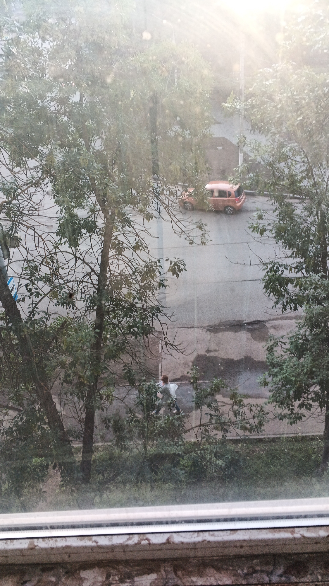 An aerial view, from a high window or balcony, looks down upon a street in Izhevsk, Russia, during what appears to be a slightly overcast or hazy day, with sunbeams filtering through the atmosphere. The foreground is dominated by the ledge of the window, with a textured brick wall below it. Beyond the window, a leafy tree with bare branches in parts obscures the view of the street. On the street, an orange, hatchback car is parked. Further down the road, a person in light-colored clothing is walking. The street itself appears to be asphalt, with some wet patches and cracks visible. The overall scene suggests a residential area within the city.