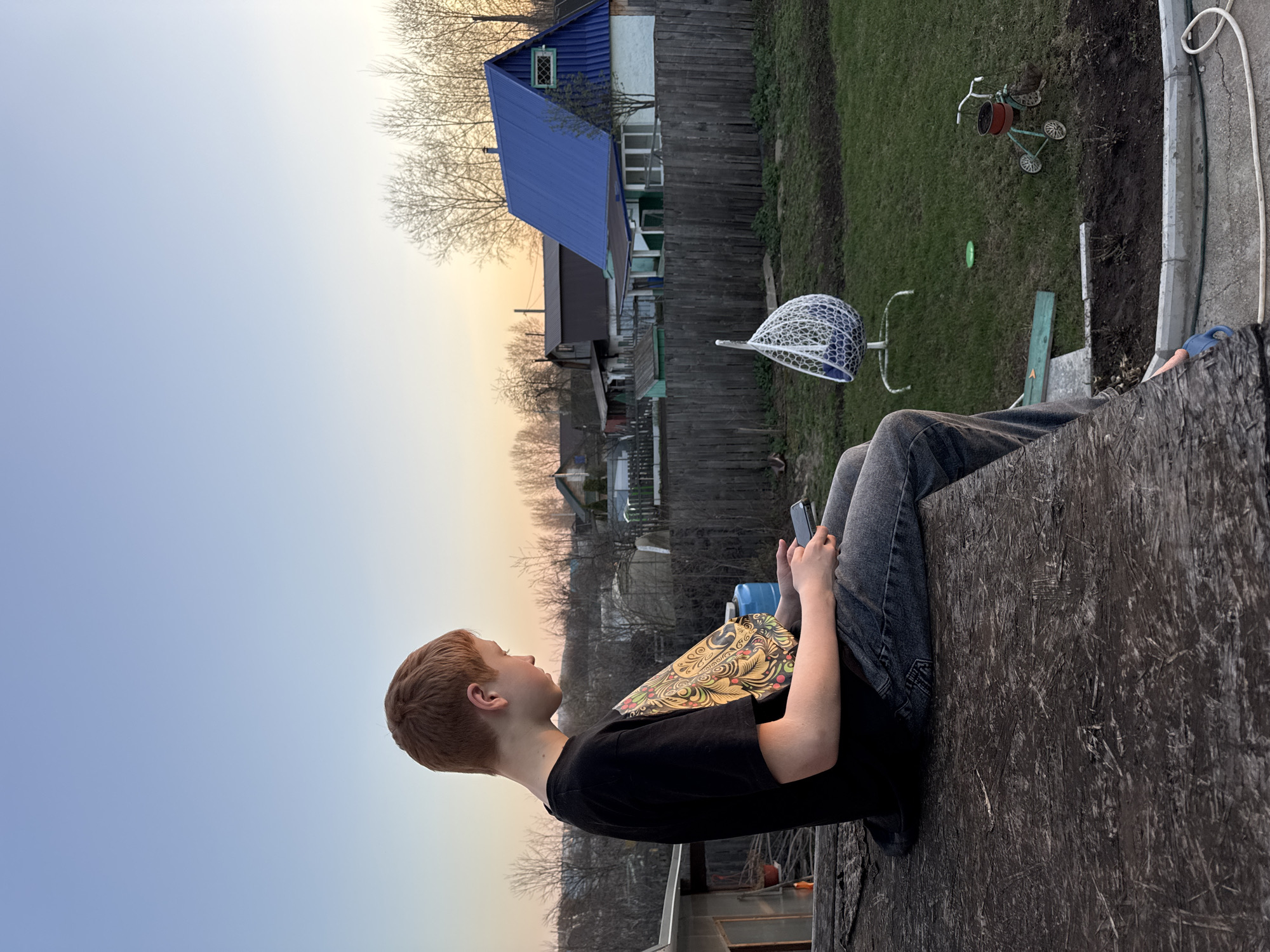 A red-haired boy sits on a dark, textured wooden surface, possibly a rooftop, overlooking a backyard in Chaikovsky, Russia. He wears dark jeans and a black t-shirt, holds a smartphone in one hand and a colorful book in the other, and gazes towards the distant trees and houses. In the backyard, a white net hangs near a green lawn with a small tricycle and a green disc. A blue-roofed house is visible beyond a wooden fence. The sky is a gradient of blue and orange, suggesting either sunrise or sunset.