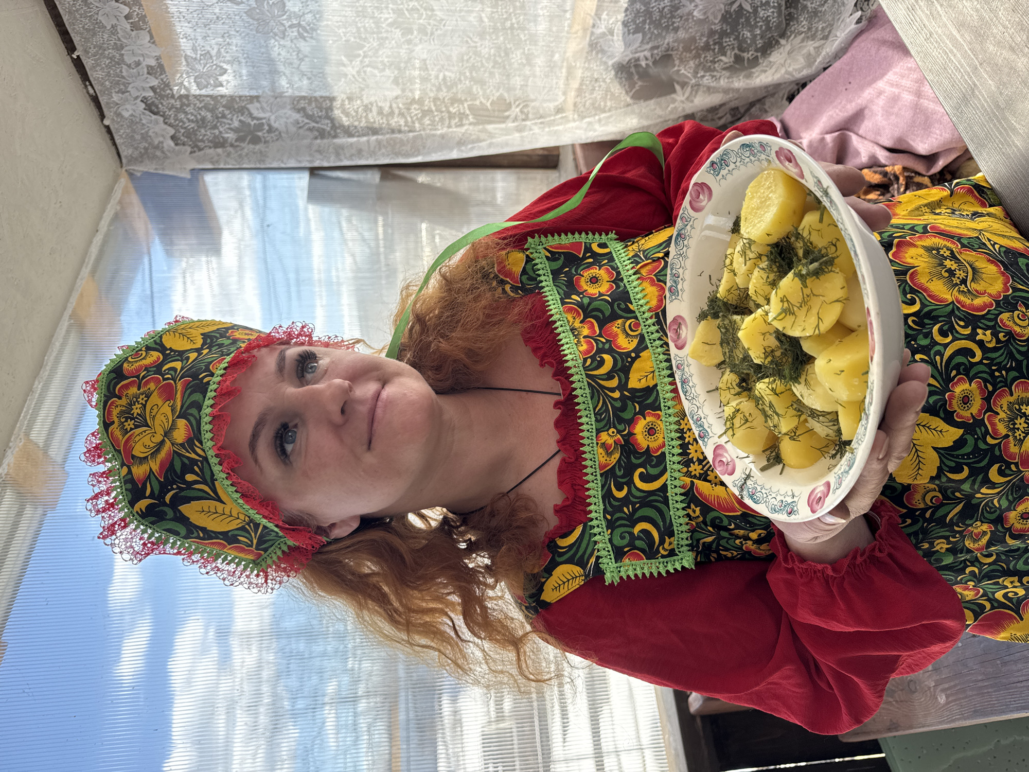A woman with red hair is holding a bowl of boiled potatoes with dill in what appears to be a greenhouse. She is wearing a traditional Russian dress with red sleeves and a floral pattern. She is also wearing a floral hat with red lace trim. The greenhouse has translucent panels and white lace curtains in the background. The photo was likely taken in the city of Chaikovsky, Russia.