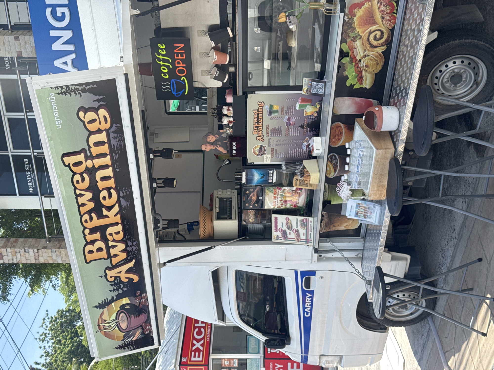 A white food truck, branded "BREWED AWAKENING," is parked outdoors on a paved surface. The truck features a large sign on its side with the name of the establishment and an illustration of a steaming coffee cup amidst a forest landscape. Above the service counter, a "COFFEE OPEN" sign with neon lights is visible. Various drinks, food items, and promotional materials are displayed on shelves and countertops, including a menu board, cups of coffee, and bottles of water. Two folding stools are placed in front of the truck. In the background, buildings and trees are visible under a clear sky. The scene appears to be a daytime street-side setting, likely in an urban area.