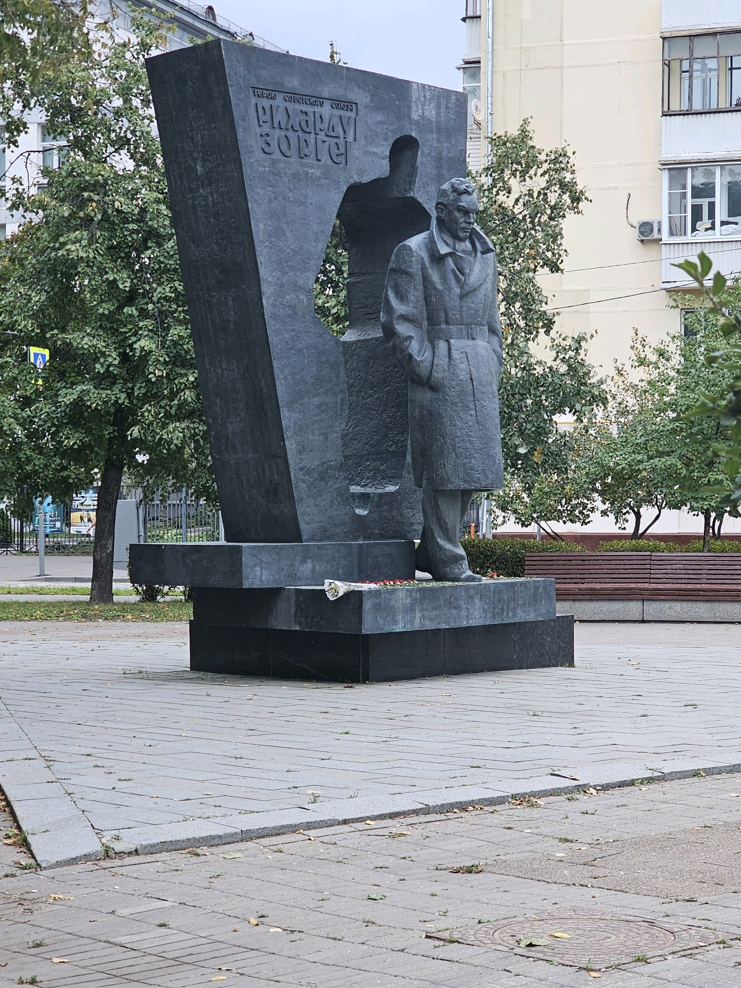 The image shows a monument to Hero of the Soviet Union, Richard Sorge, located in Moscow, Russia. The monument is made of dark stone and features a large, abstract sculptural element with an opening, behind which stands a statue of Sorge in a trench coat. In front of the monument, there is a base with some fallen petals. The monument is situated in an urban environment with a paved plaza, trees, buildings, and a street visible in the background. The overall atmosphere is somber and monumental.