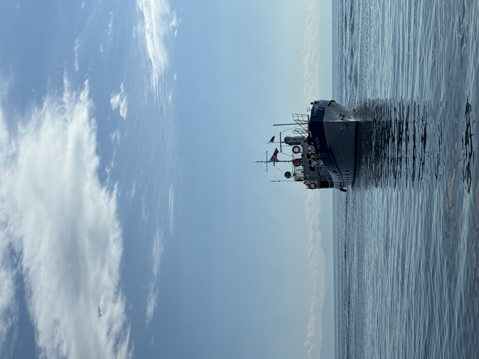 A forward-facing view of a small, light-blue and white passenger boat named "РОССИЯ" is captured on a calm, blue body of water. The boat is at a medium distance, with its bow pointed directly towards the viewer. The water is mostly calm, with subtle ripples creating a textured surface and reflecting the sky. In the distance, a hazy, mountainous shoreline can be seen under a bright blue sky with scattered white clouds. The boat has several life preservers visible on its exterior and appears to have people on its deck. The scene suggests a bright, clear day, possibly in the city of Listvyanka, Russia, given the style of the boat and the surrounding natural landscape, which is consistent with the area.